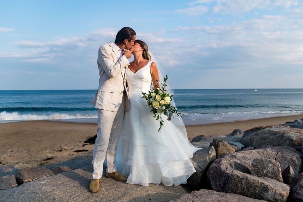 A couple in wedding attire embrace on a rocky beach with waves and blue sky in the background.