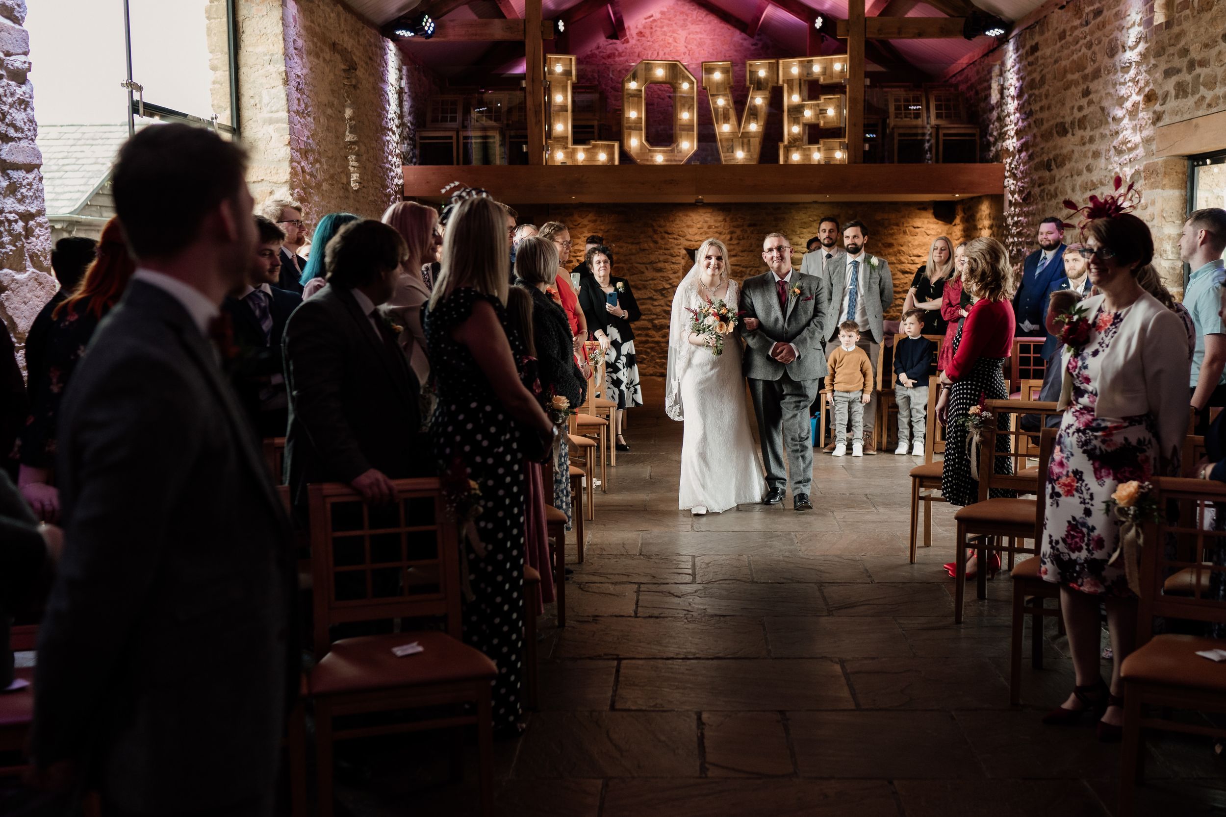 Wedding ceremony in a rustic Dodford Manor with illuminated LOVE sign and guests seated in rows.