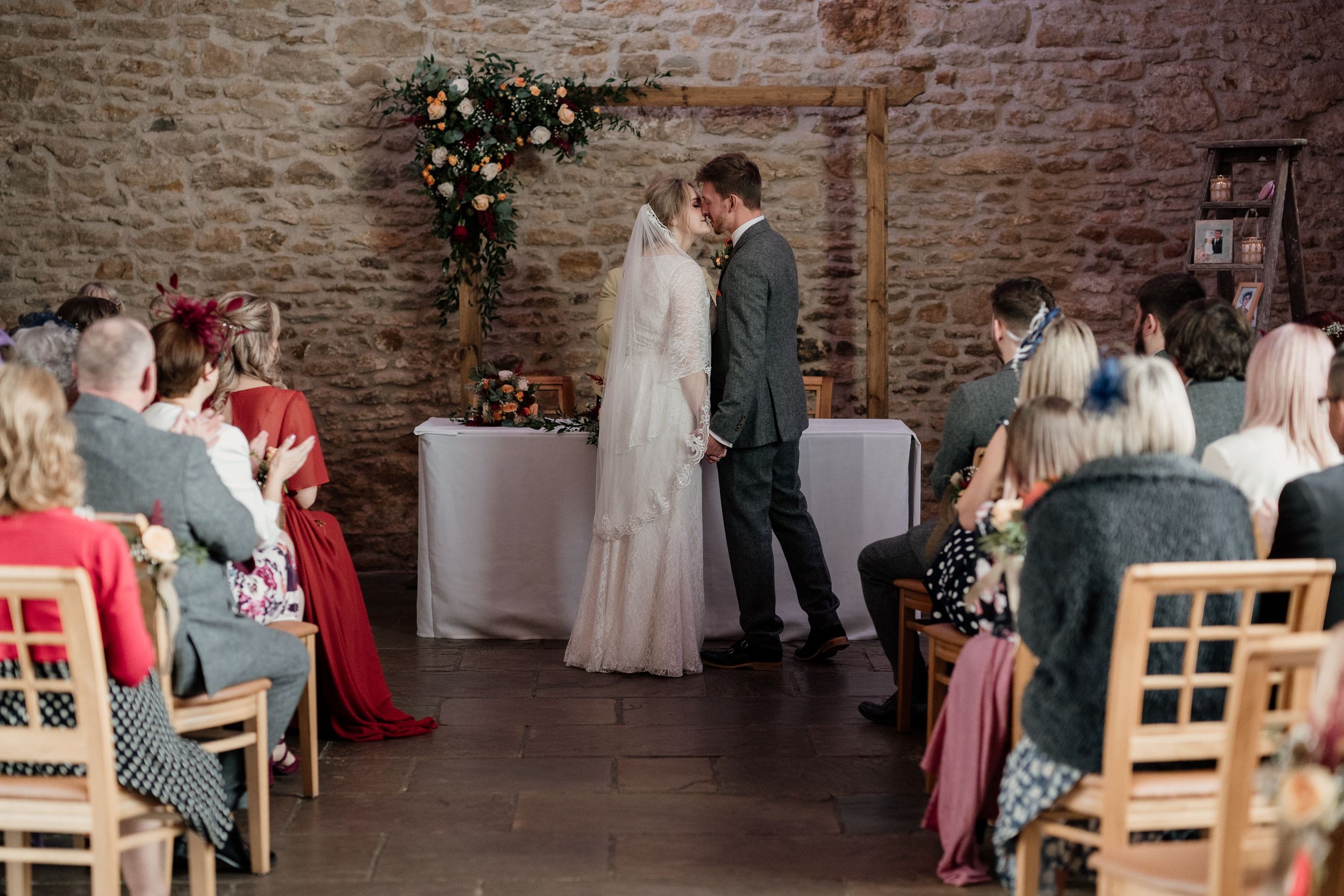 Intimate wedding ceremony inside a rustic stone barn at Dodford Manor with wooden chairs and floral decorations.