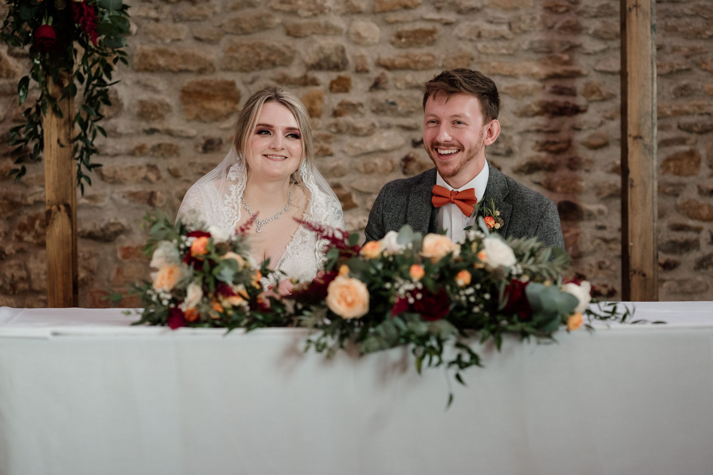 Couple seated at wedding head table with lush floral arrangements and greenery.