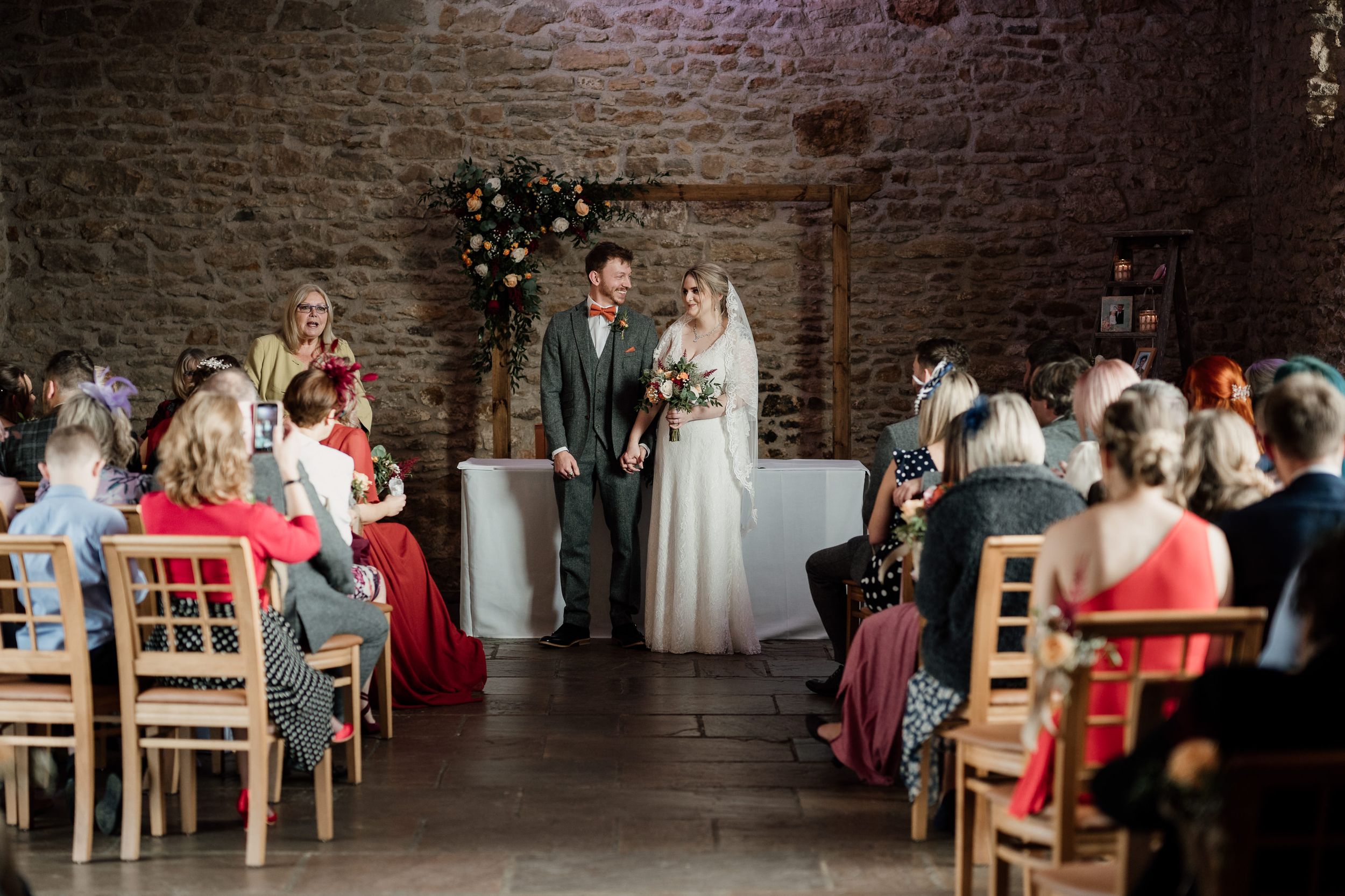 Wedding ceremony in progress inside rustic stone venue with wooden chairs and floral arch, Dodford Manor.