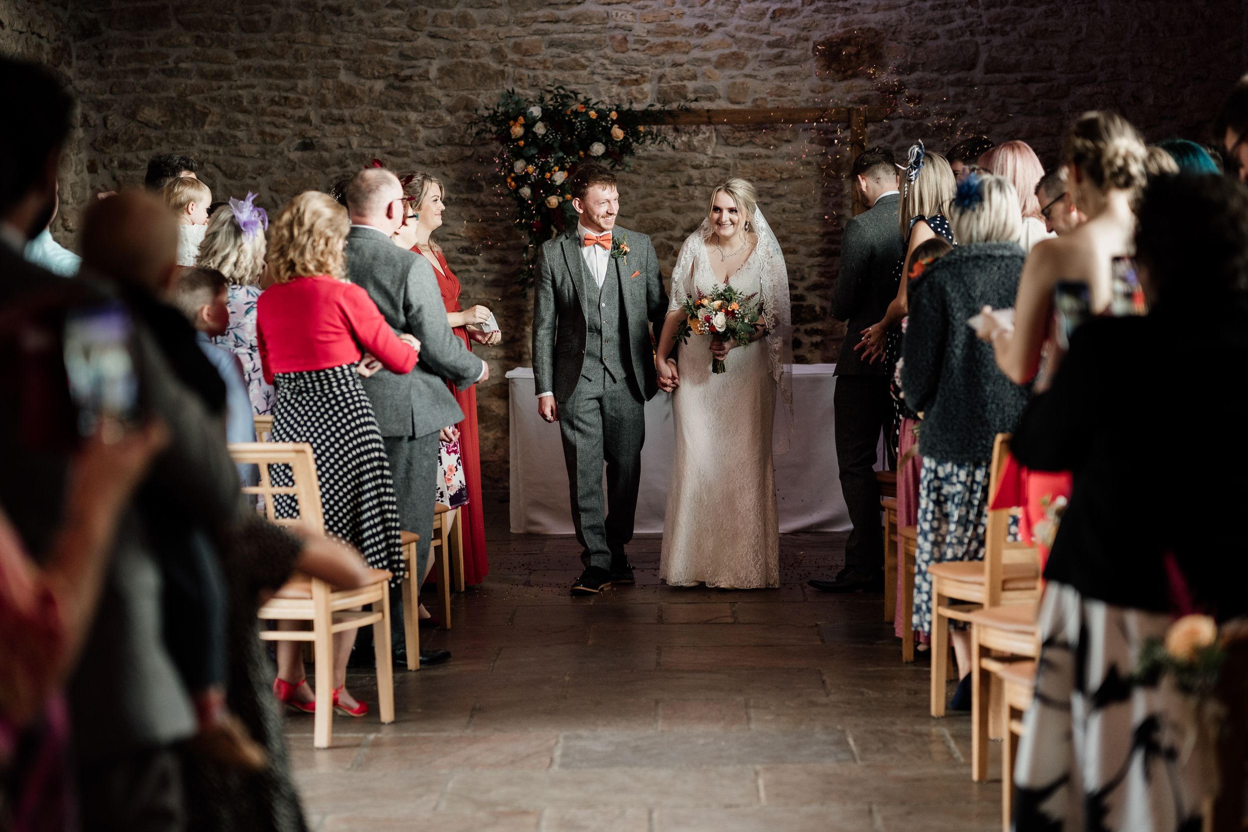 Wedding processional in stone-walled venue with guests seated on wooden chairs.