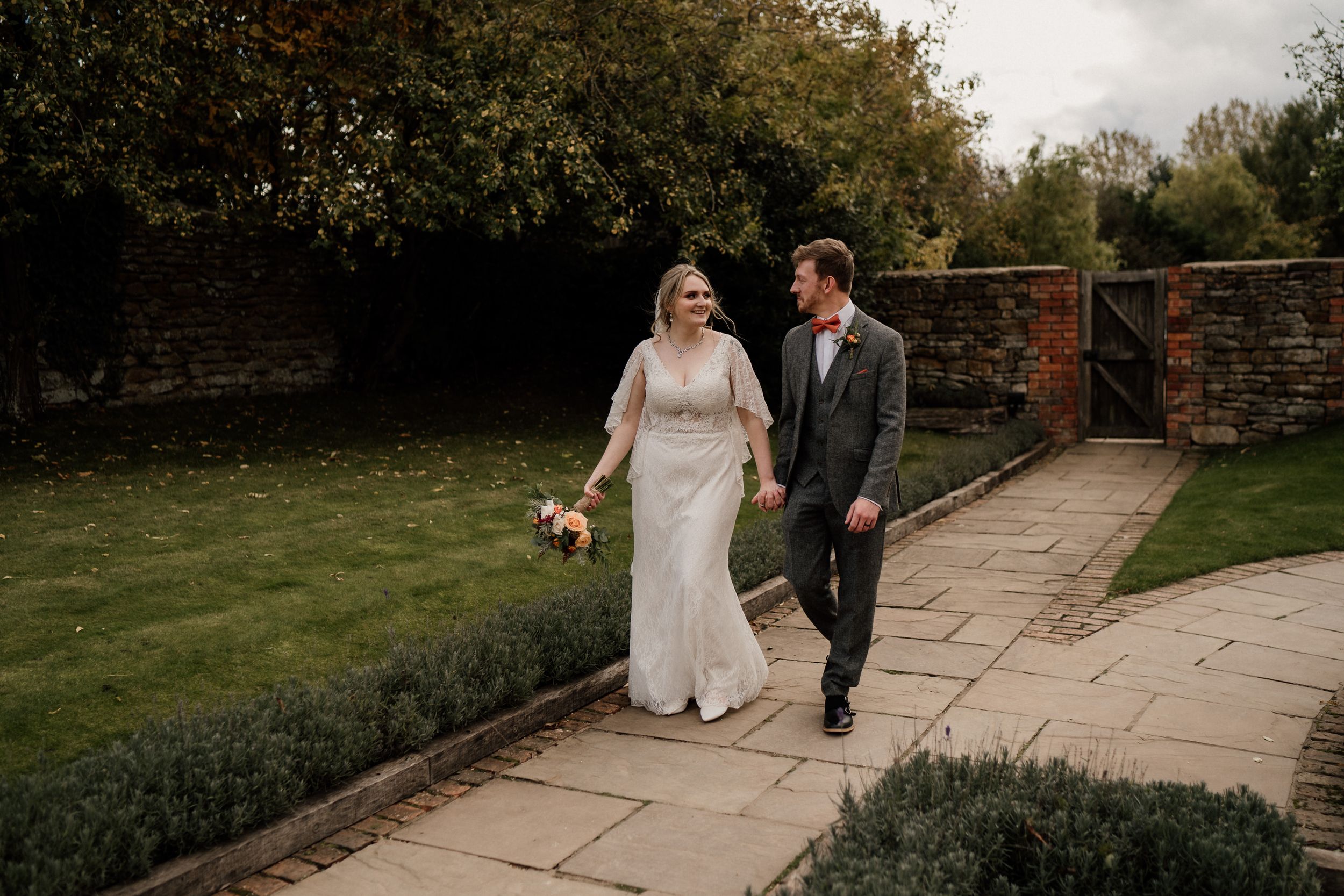 Newlyweds walking together along stone garden path holding autumn bouquet.