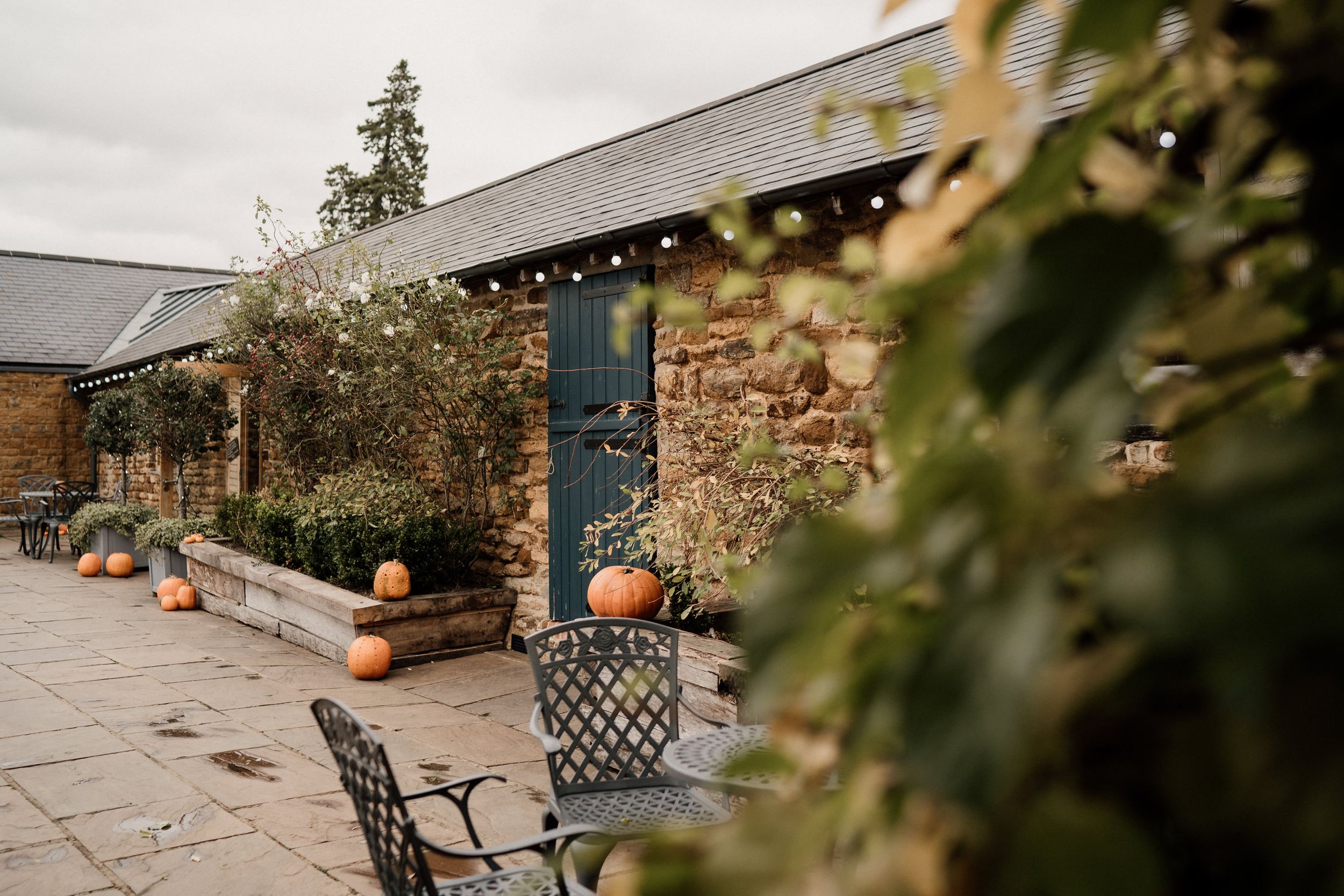 Rustic stone building exterior of Dodford Manor with pumpkins and outdoor seating on a patio.