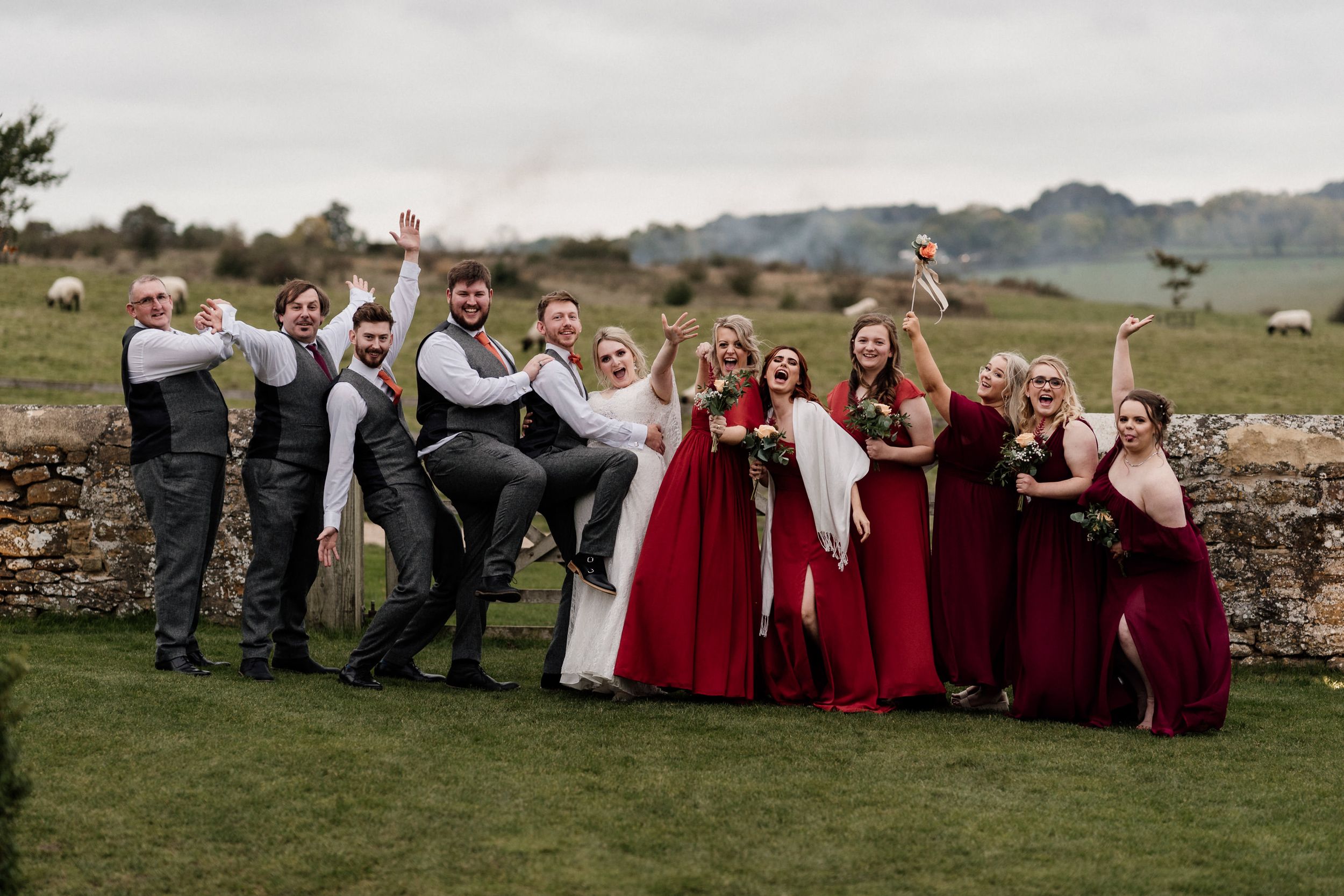 Wedding party in formal attire poses playfully against a stone wall in a countryside setting at Dodford Manor Barns.