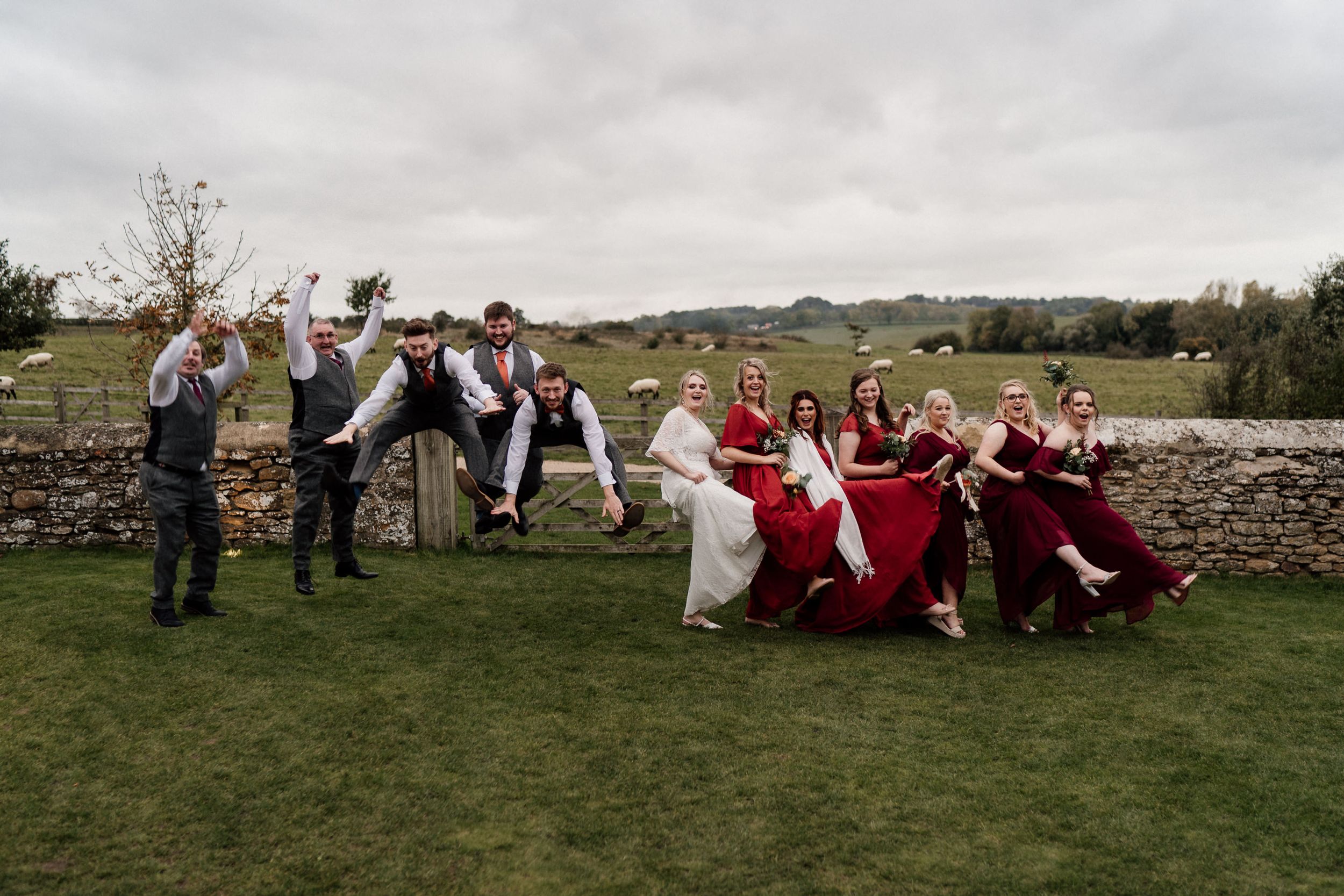 Bridal party splits into two groups striking dynamic poses on grassy lawn by stone wall.