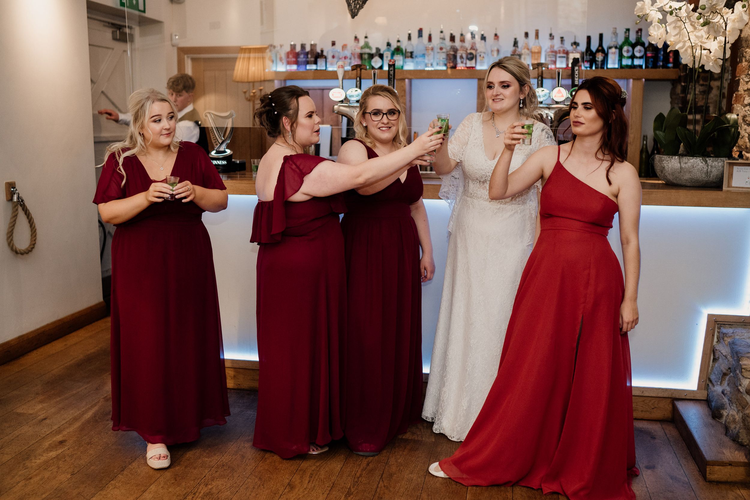 Bridal party in burgundy dresses gathers at an elegant indoor bar area.