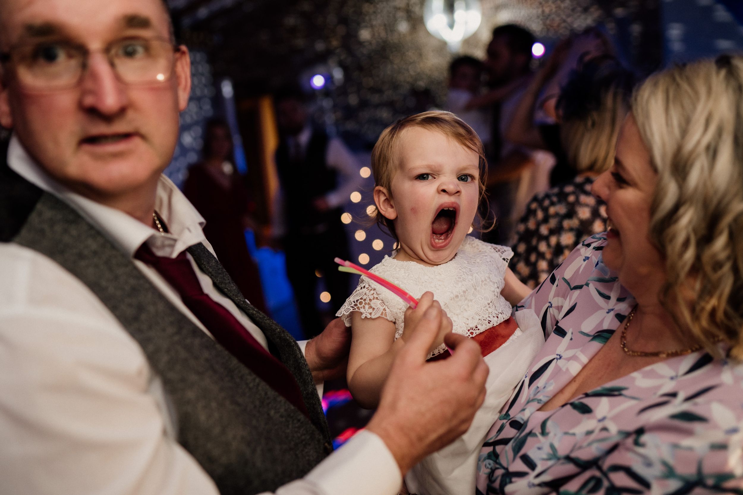 A yawning baby in white dress attends a wedding celebration.