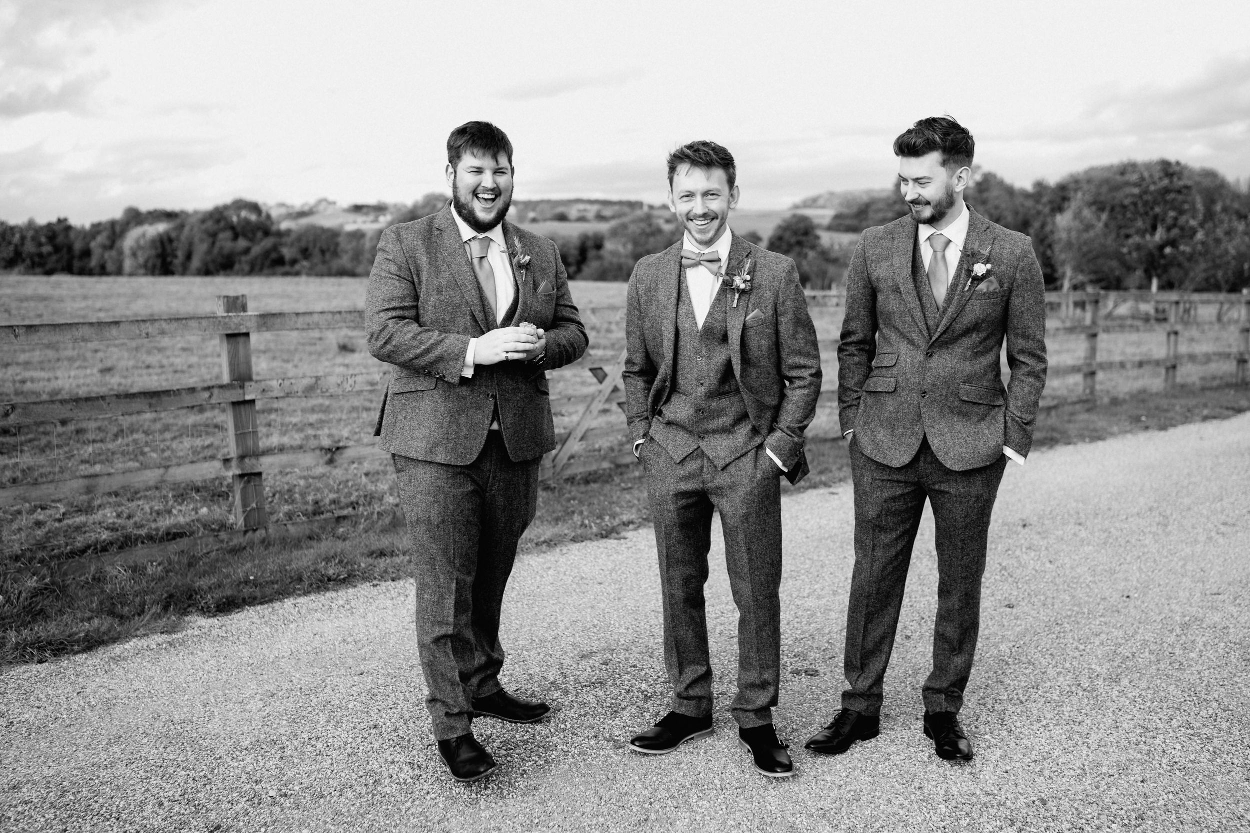 Three groomsmen in suits posing on a rural path in black and white.