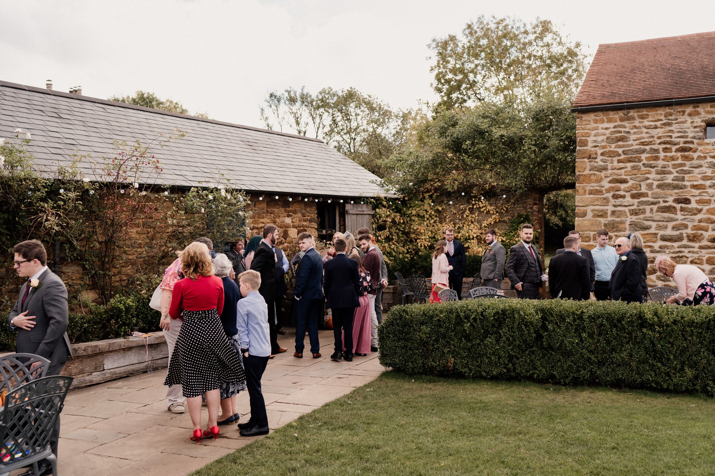 Wedding guests mingling in an outdoor courtyard at Dodford Manor with stone buildings and manicured gardens.