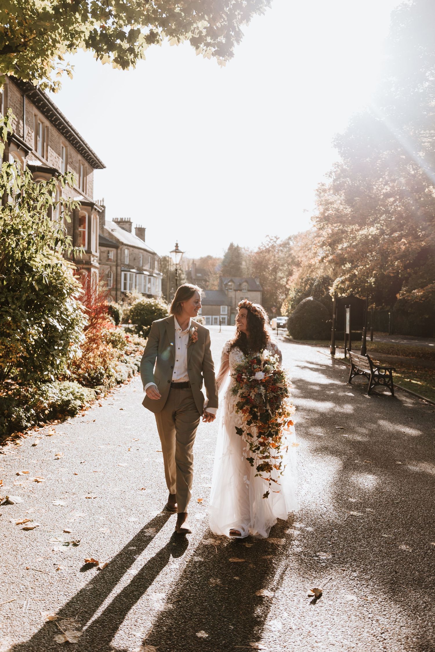 A couple strolls hand in hand down a sunlit street lined with autumn trees casting long shadows on a romantic wedding day.