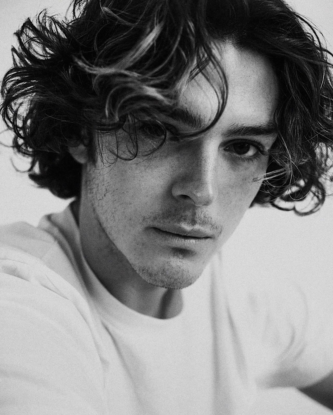 Black and white portrait of a person with tousled dark curly hair wearing a white t-shirt against a plain background.