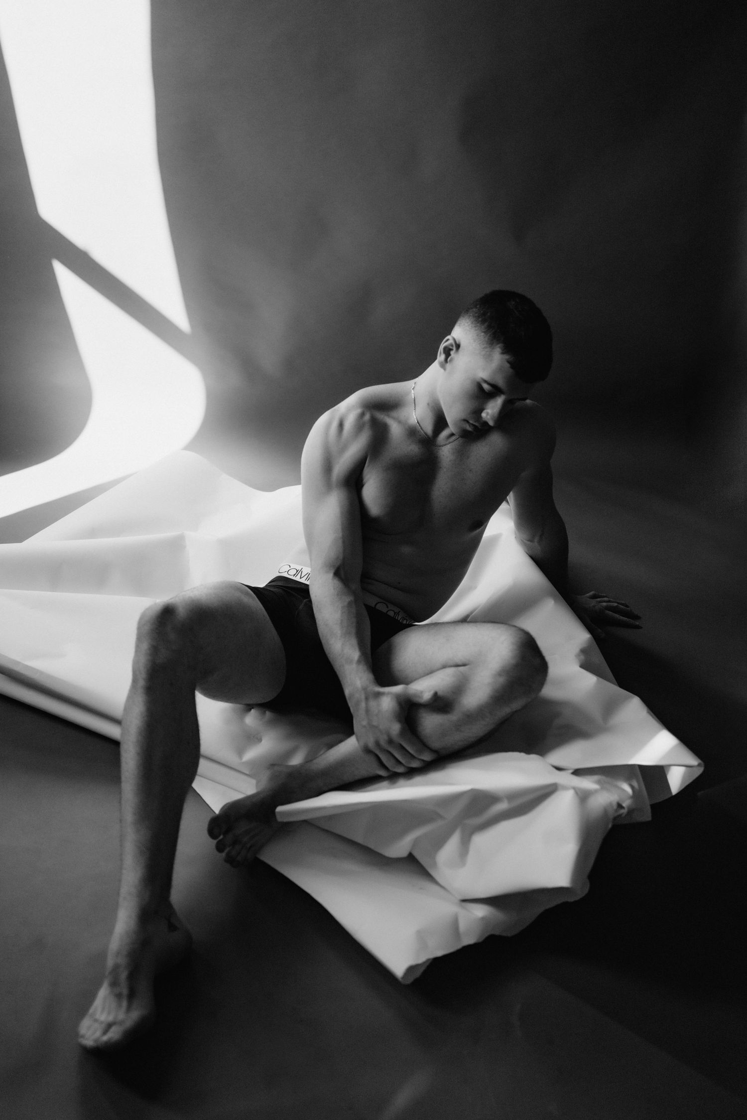 Artistic black and white photo of shirtless athletic figure posed on crumpled paper in dramatic lighting.