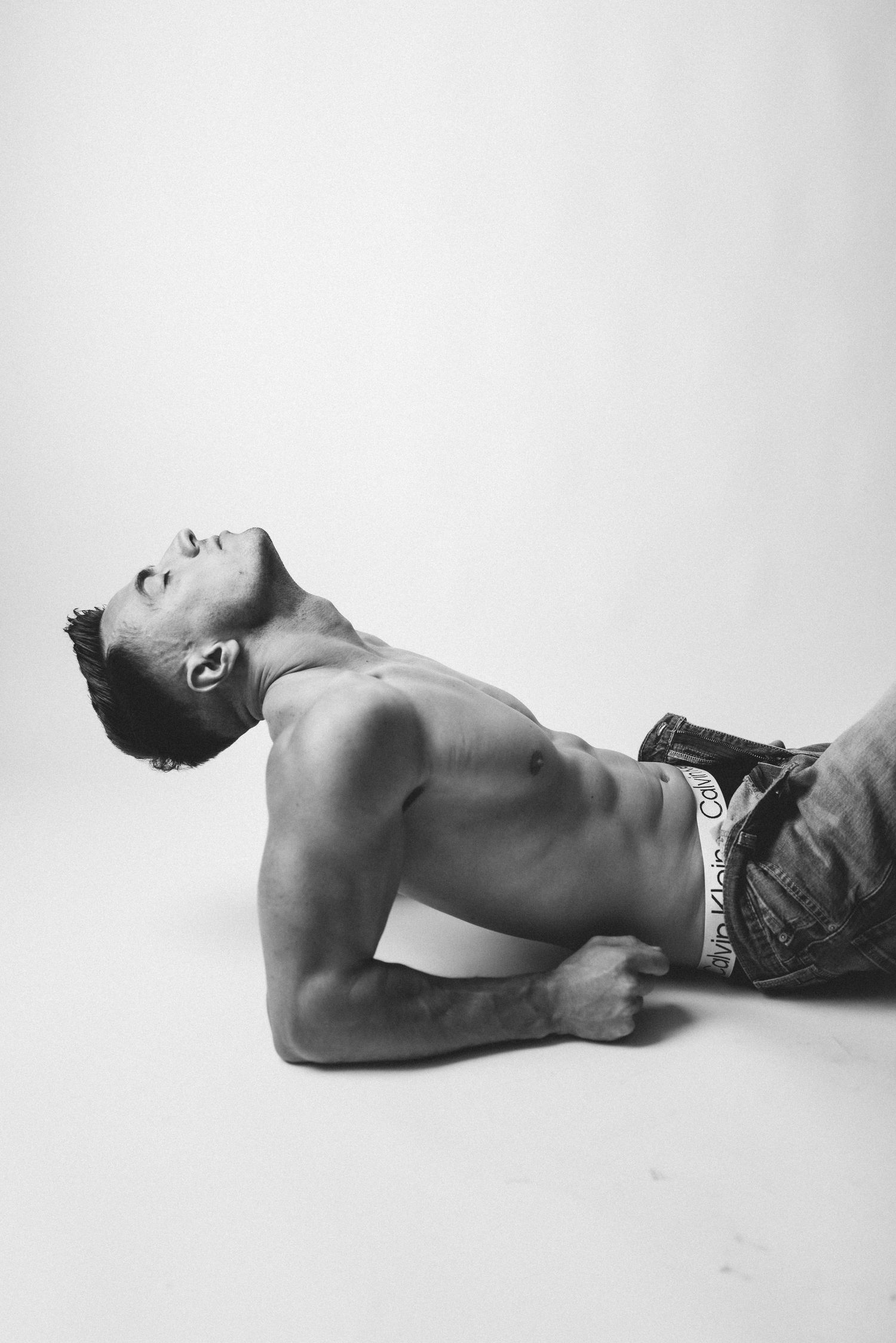 Black and white artistic photograph of shirtless athletic model in jeans posing against white background.