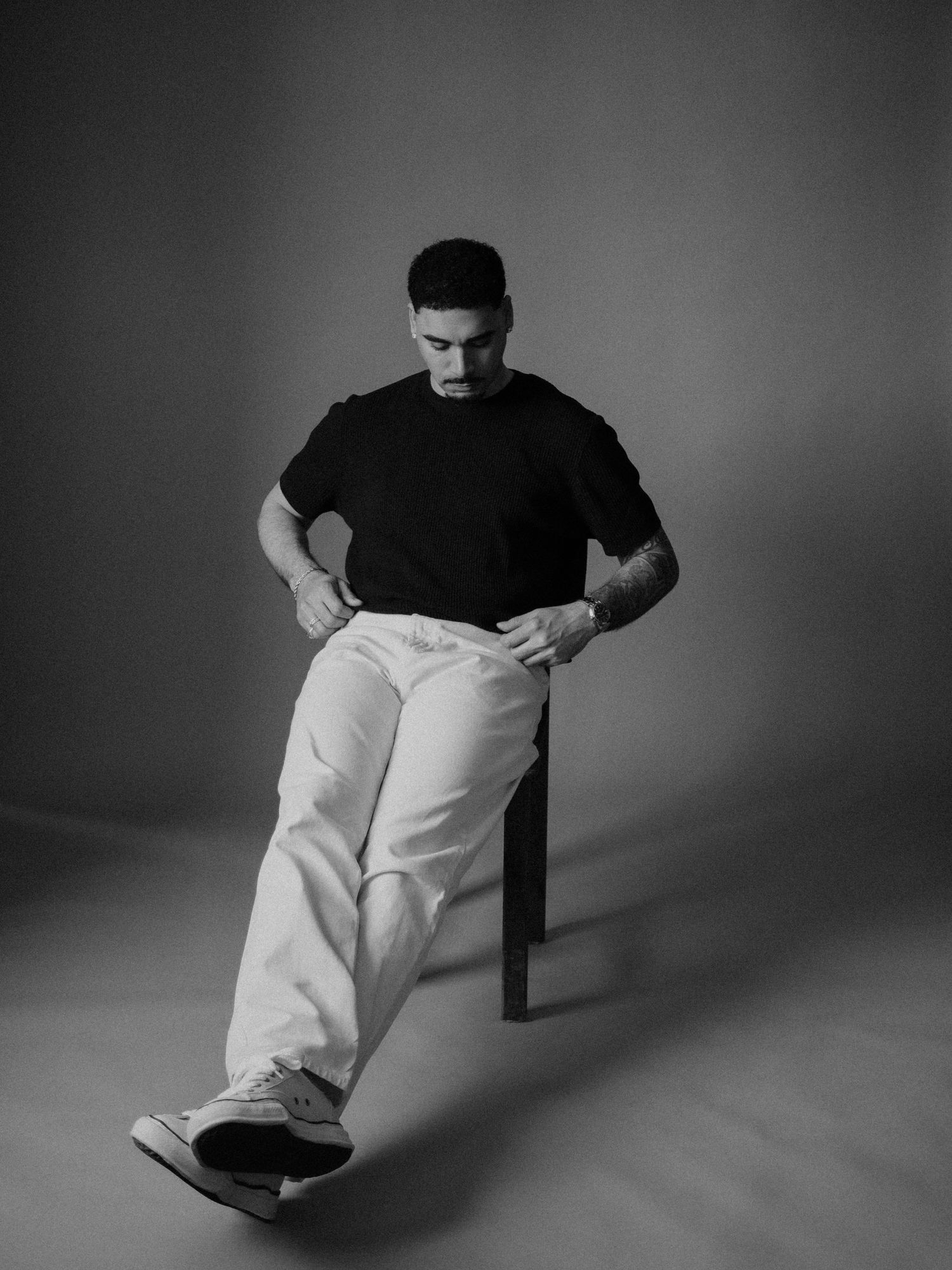 Artistic black and white portrait of a person in a black t-shirt and white pants sitting casually on a chair against a gray backdrop.