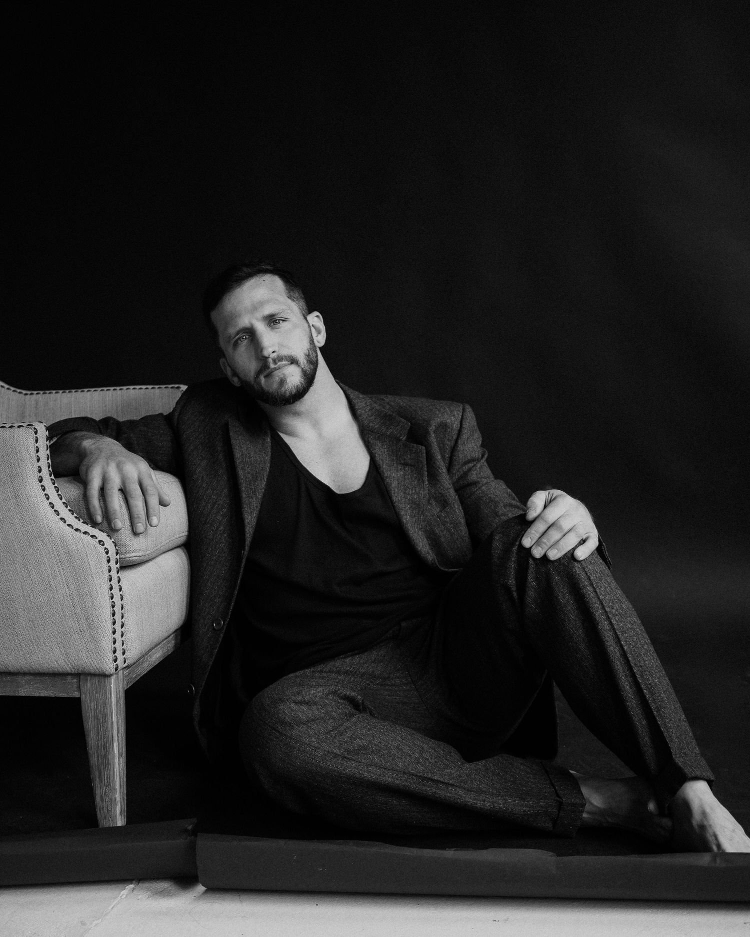A black and white portrait of a bearded man in a dark suit and v-neck shirt lounging against an upholstered chair.