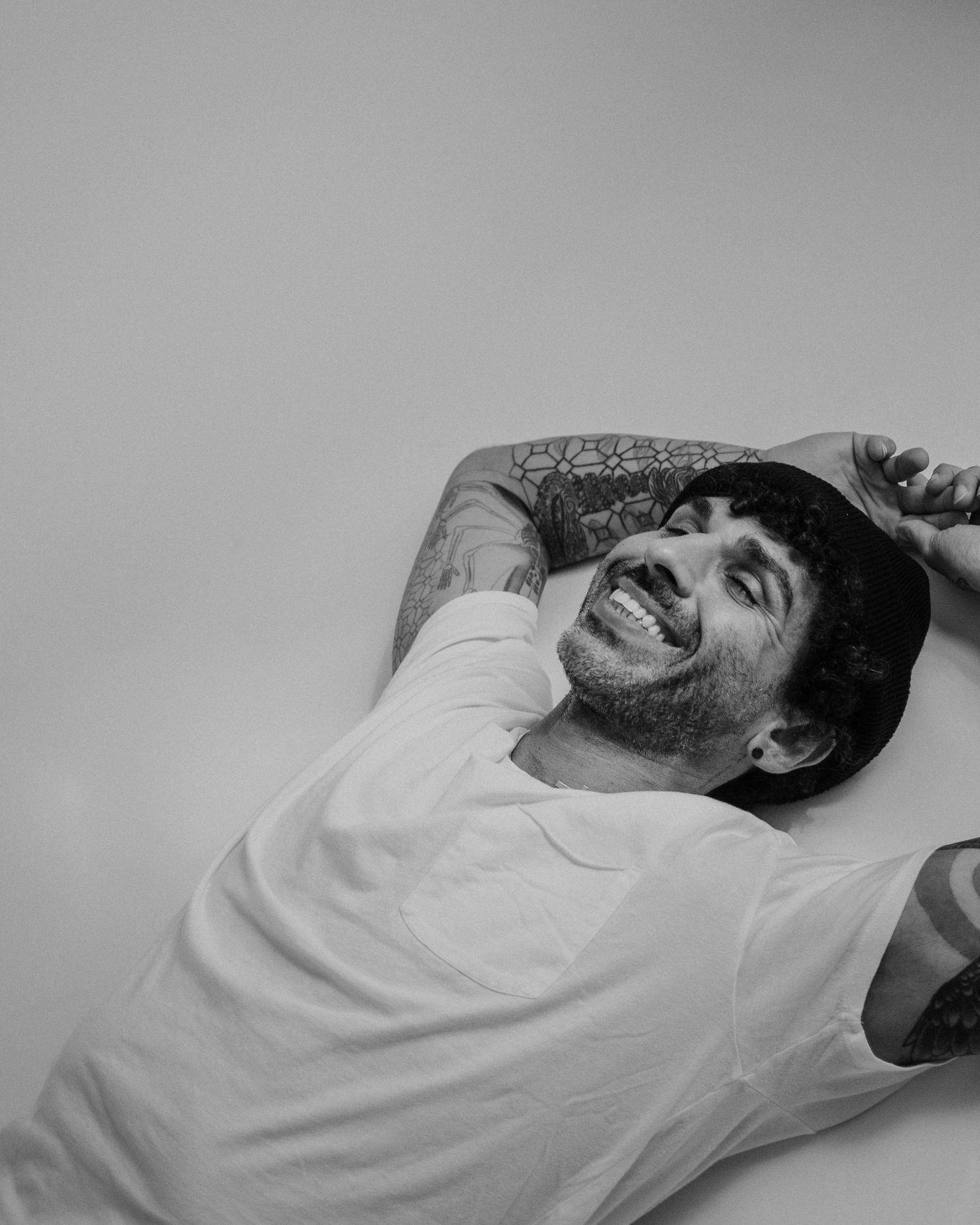 A smiling person in a white t-shirt lies back on a couch with tattoos visible on their arm.