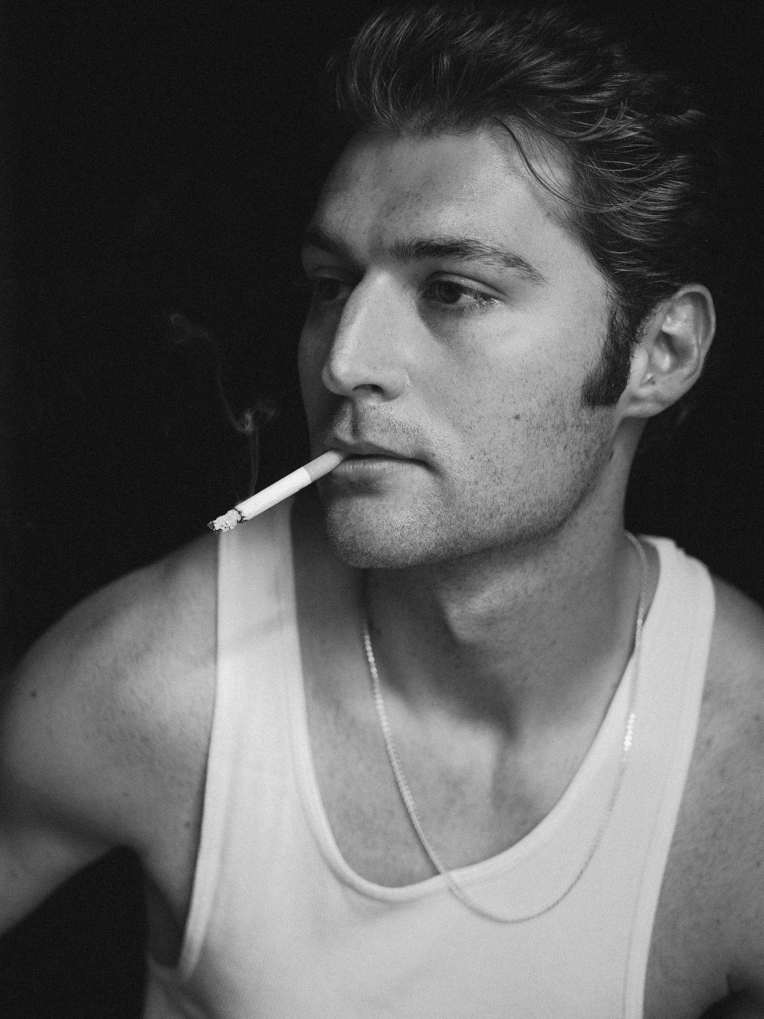 Black and white portrait of a person in a white tank top smoking a cigarette against a dark background.