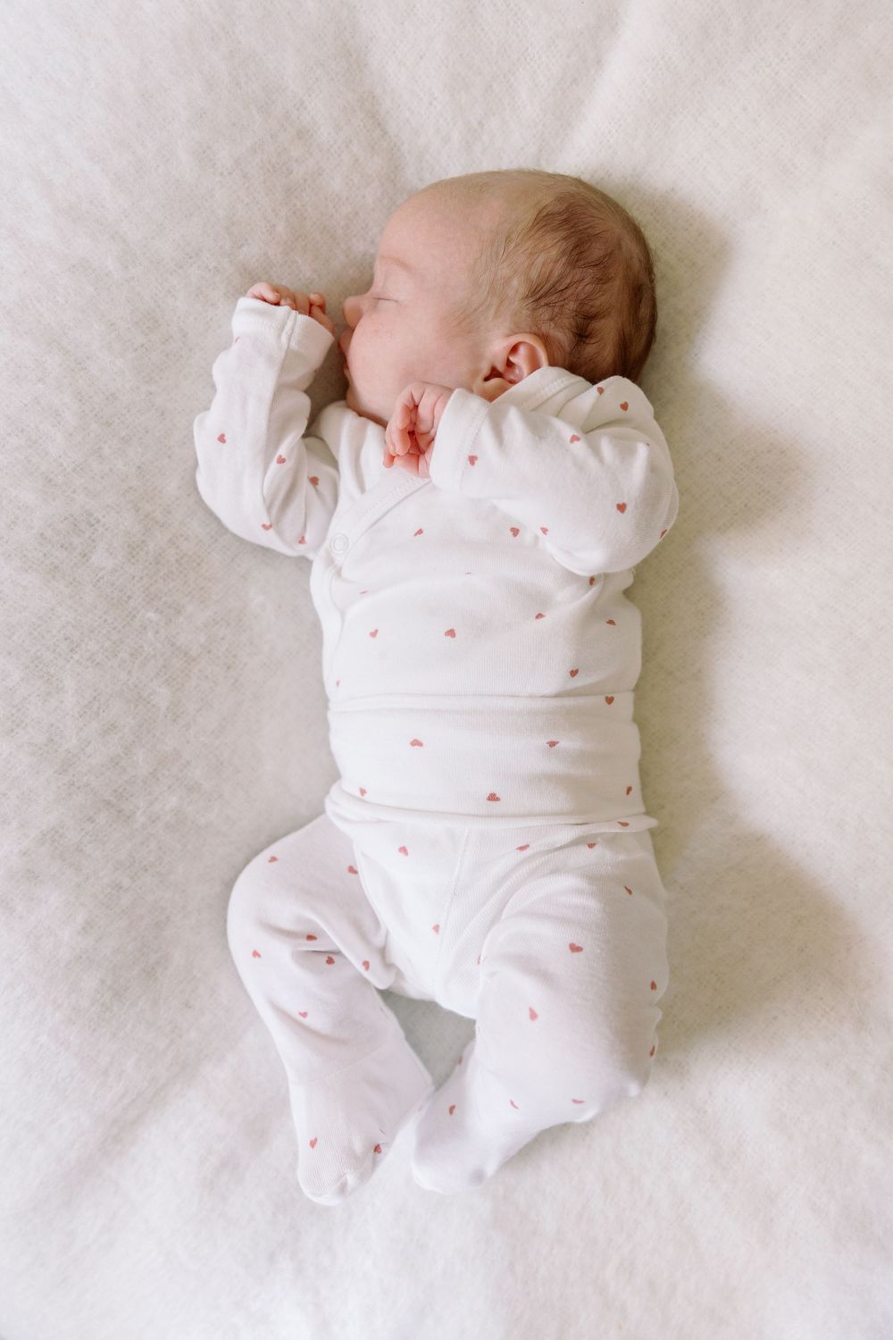 Sleeping newborn wearing white onesie with tiny pink heart pattern lies peacefully on soft white bedding.