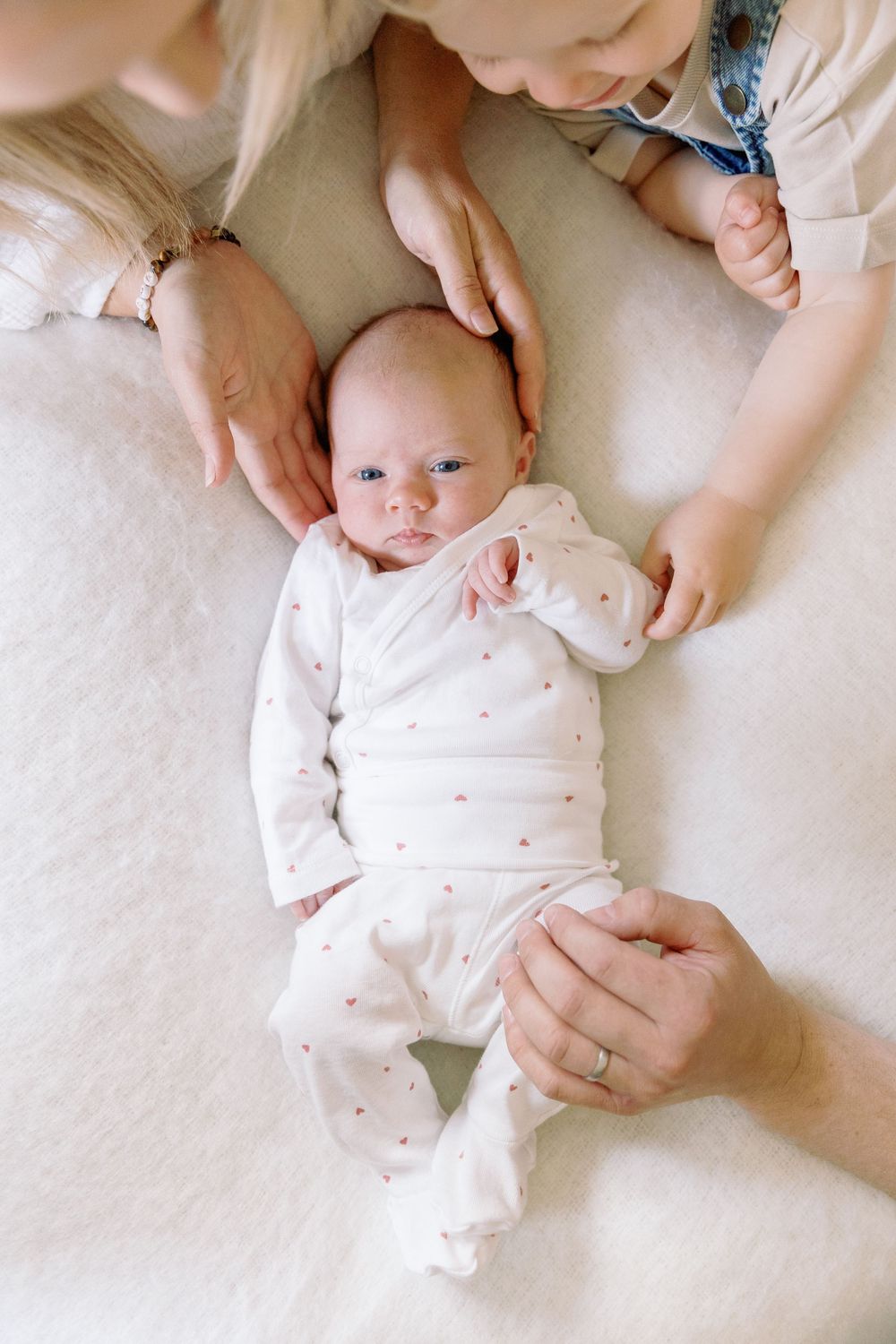 A baby in white polka dot pajamas lies on a cream colored surface while being gently cradled by caring hands.