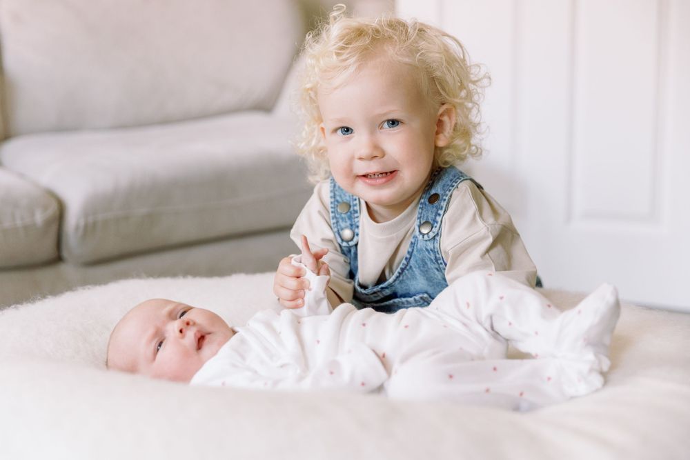 A small child with curly blonde hair wearing overalls sits next to a baby lying on a white surface.