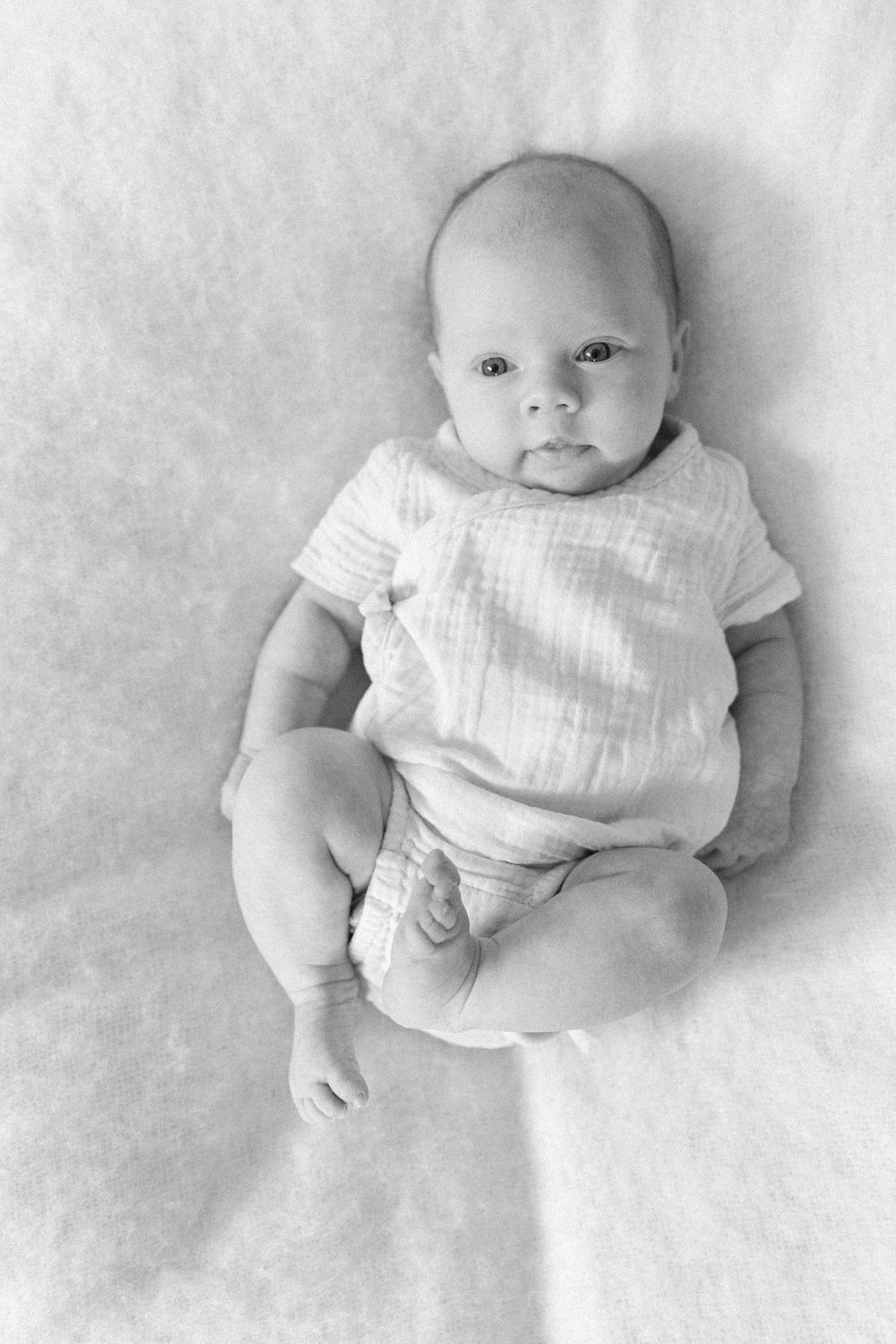 Black and white portrait of a young baby wearing a light colored onesie lying on soft fabric.