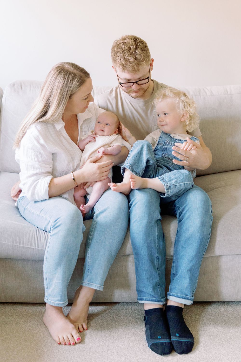 A family sits together on a light gray couch, wearing casual clothing and sharing a tender moment at home.