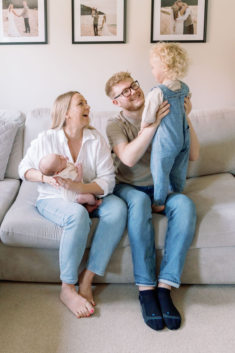 A happy family relaxes together on a grey couch with two small children, sharing joyful expressions and casual comfort.