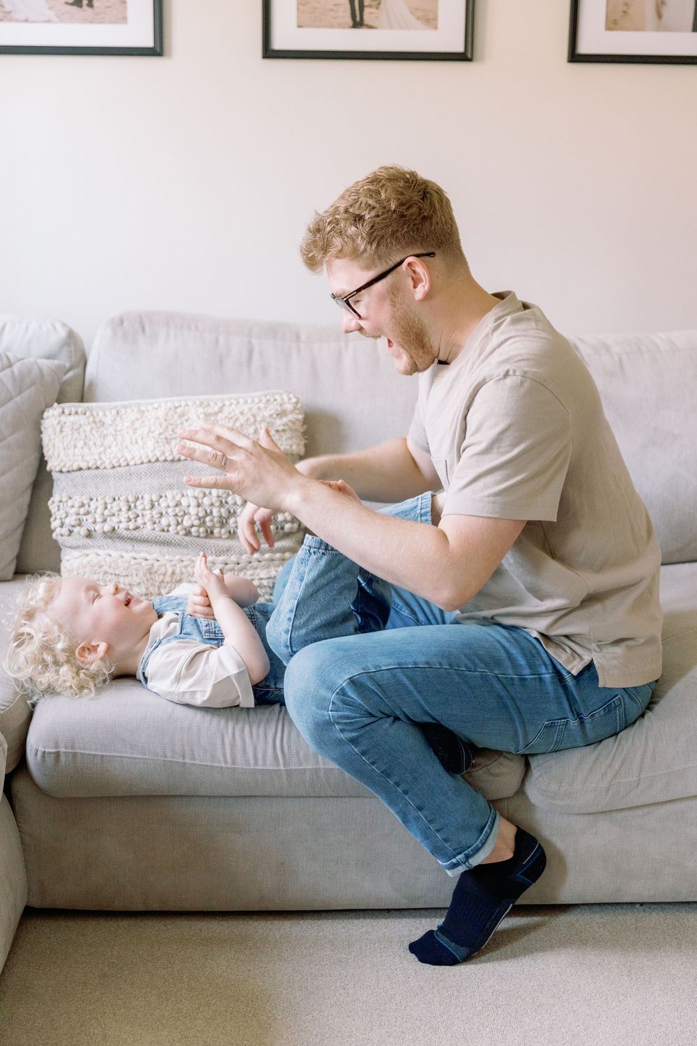 A young parent in glasses and jeans plays with a baby resting on a light gray couch with decorative pillows.