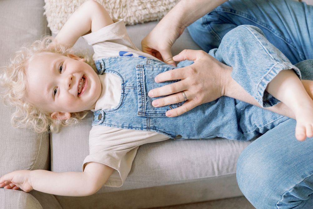 A laughing toddler in denim overalls lies on a gray couch while being tickled by adult hands.