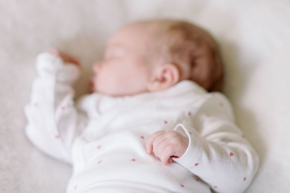 A sleeping infant wearing a soft white onesie rests peacefully against a light background.