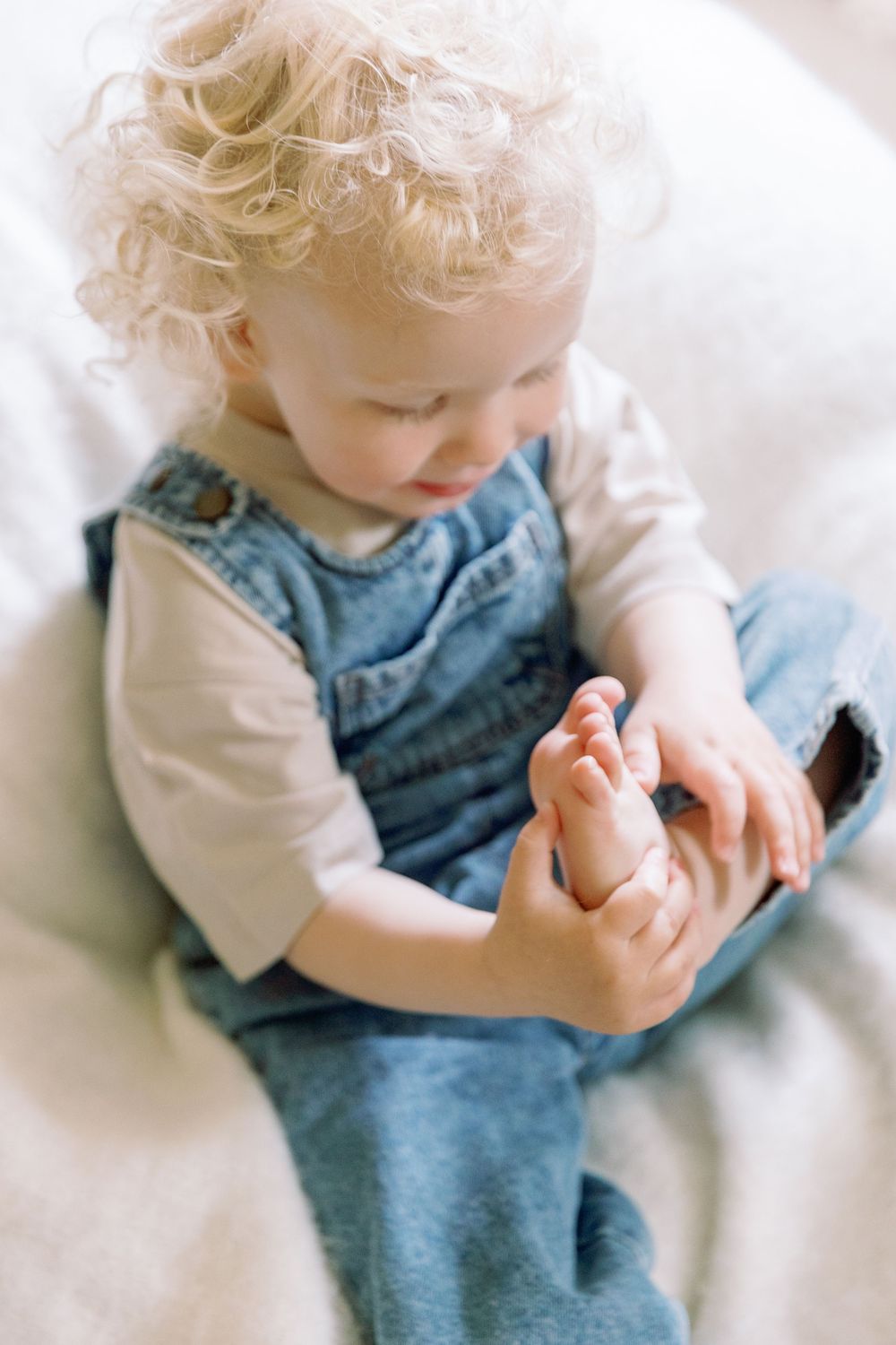 Small child in denim overalls and white shirt sits on white surface while playing with their feet.