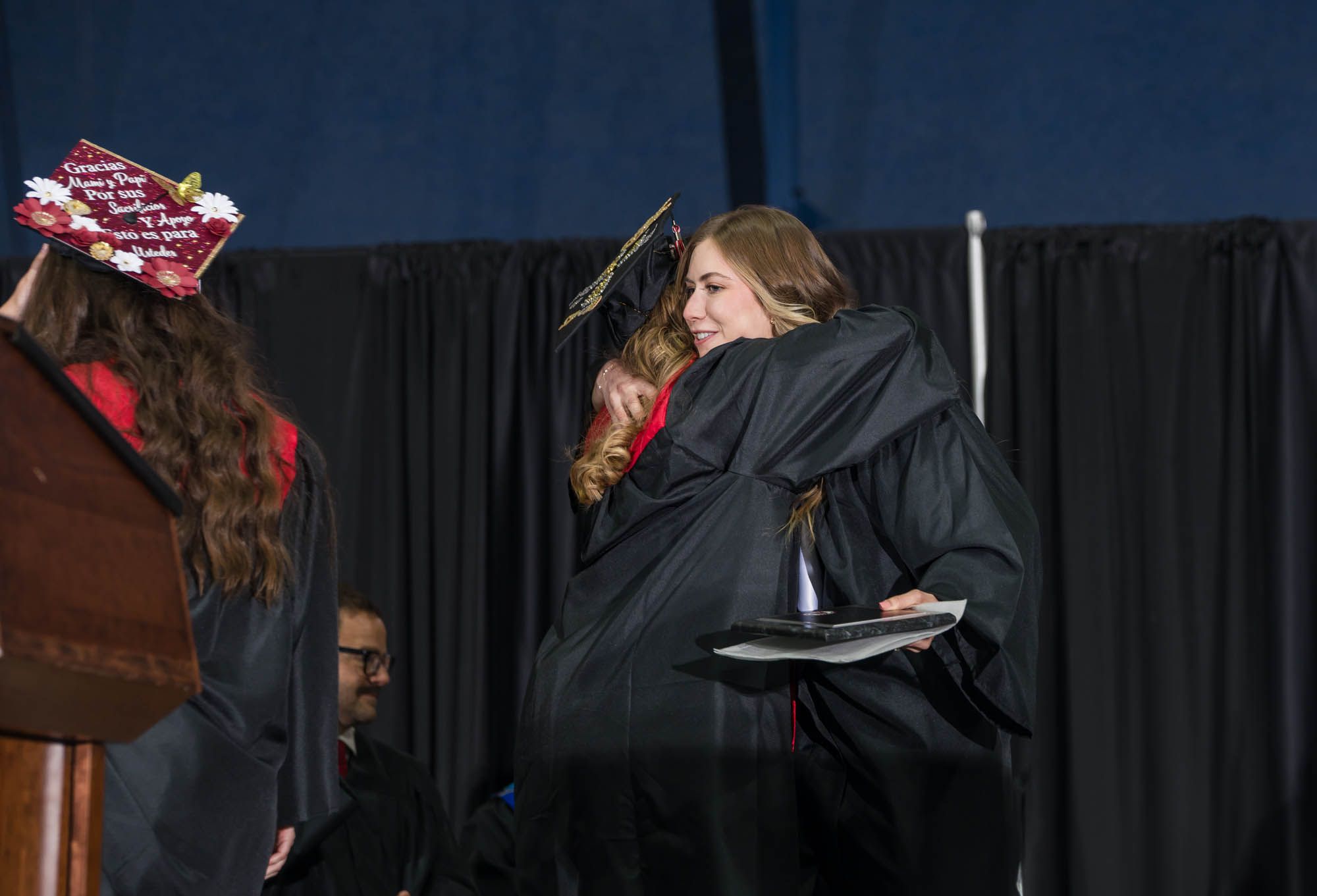 Two graduates in black caps and gowns embrace during a graduation ceremony against a dark backdrop.