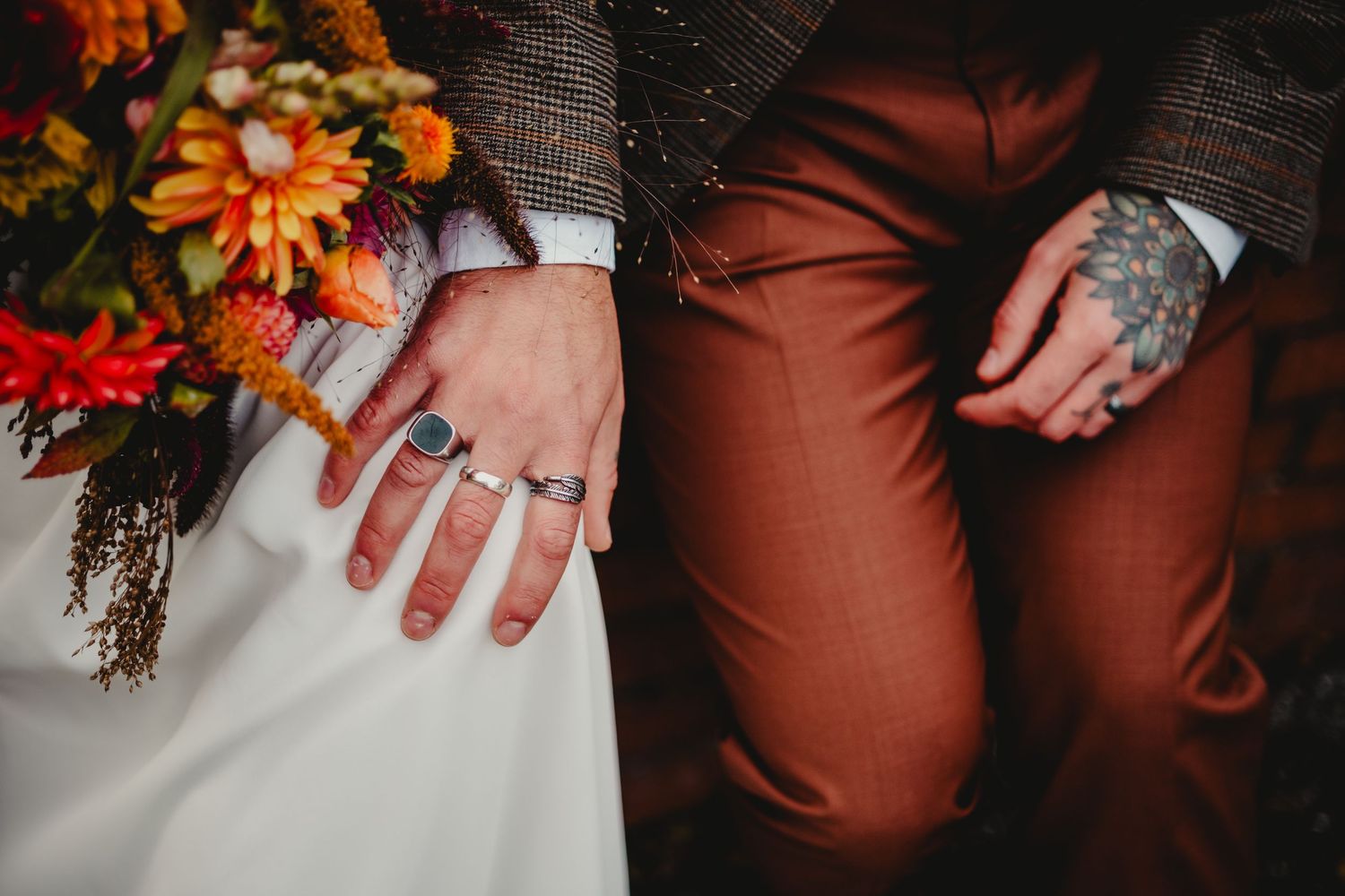 Close-up shot shows hands wearing rings next to autumn wedding bouquet with orange flowers against rustic backdrop.