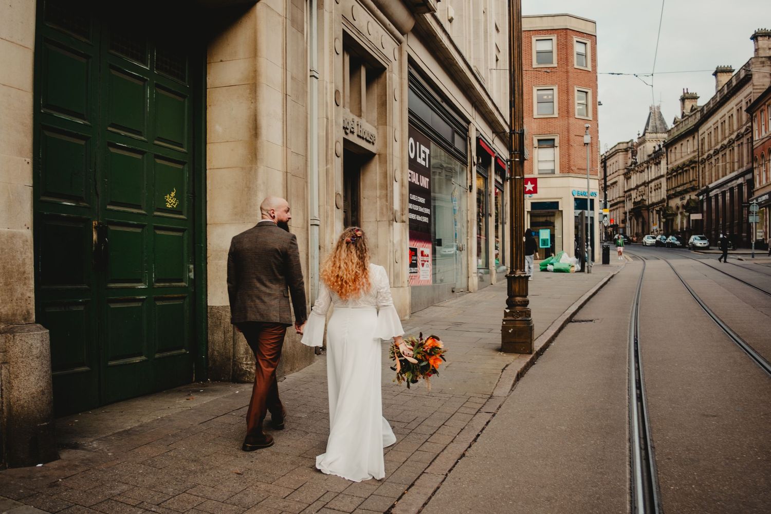 A couple walks down a historic city street with tram tracks, the person in white holding a colorful bouquet.