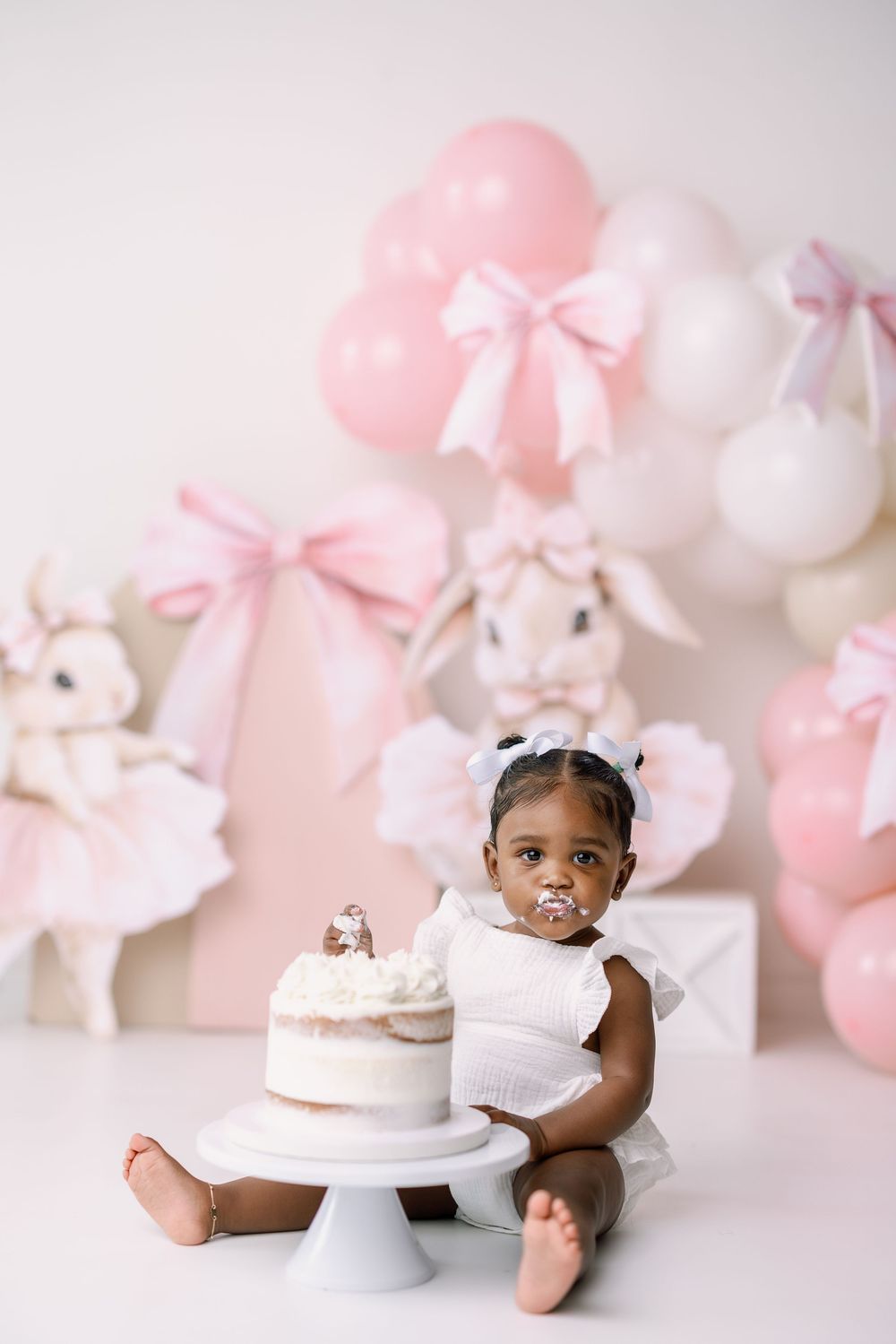 Sweet pink and white balloon decor with plush bunnies creates a delightful backdrop for a first birthday cake smash session.