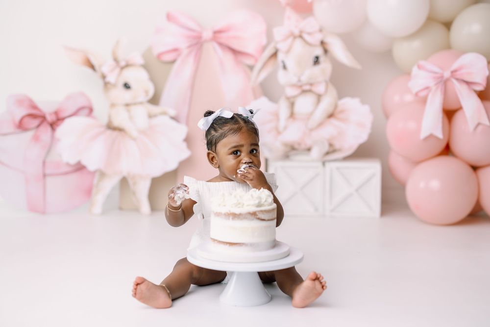 Pink baby shower decorative setup with bows, balloons, cake and festive decorations against a light background.