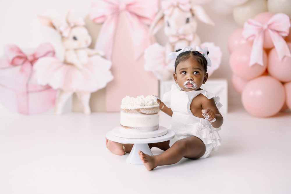 Baby in a white outfit sitting next to a cake with pink decorations and balloons in a soft pastel studio setting.