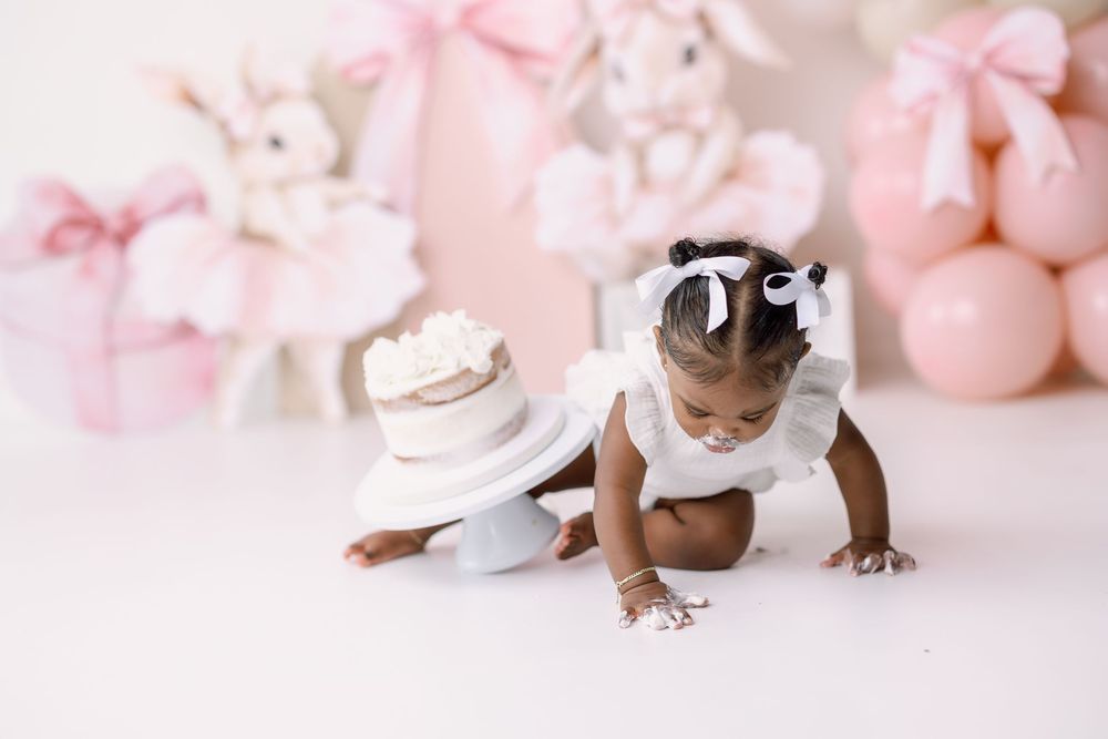 Infant in white tutu dress crawling on pink background with ballet shoes and plush toys visible in studio setting.