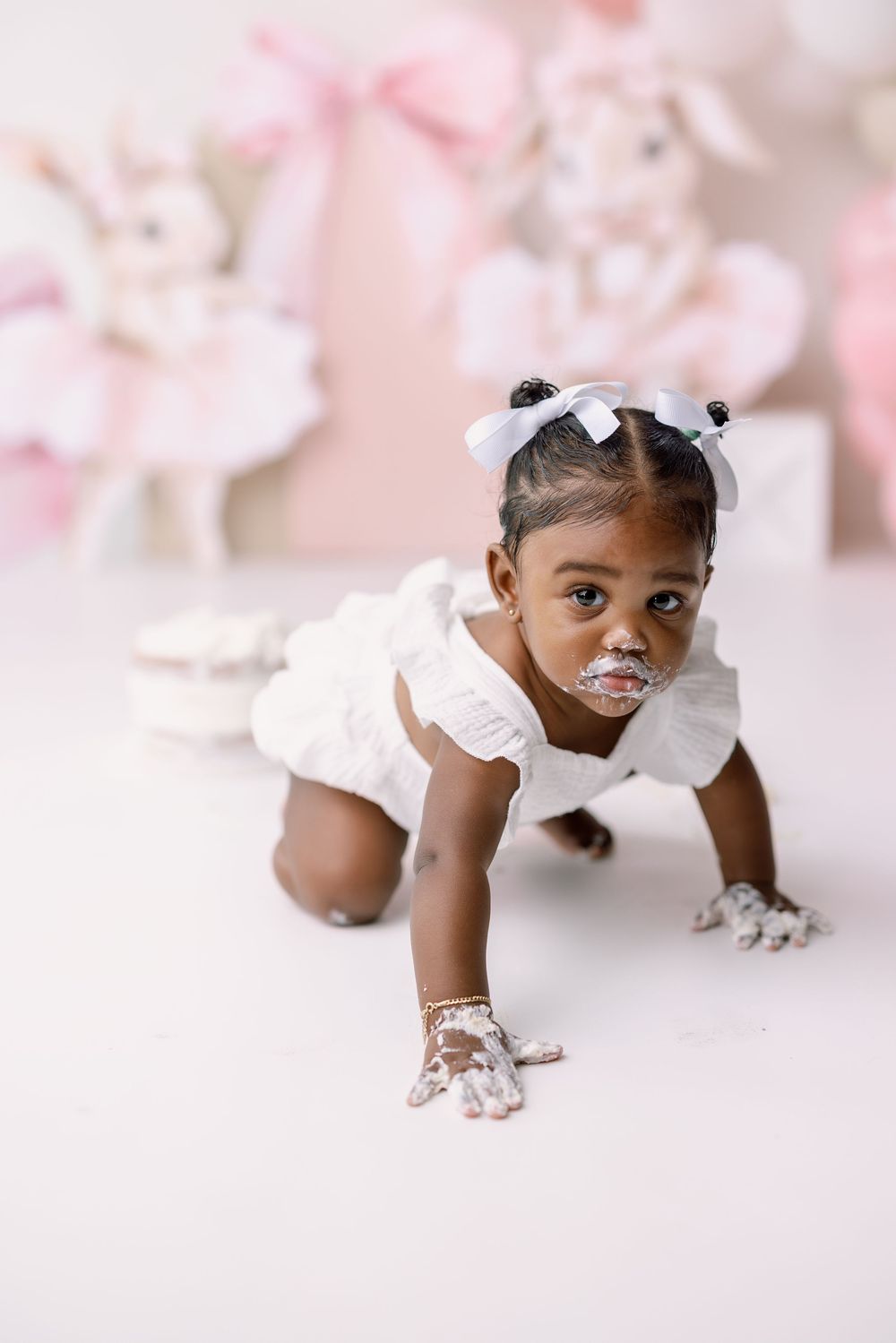 Small child wearing white ruffled dress and bows crawls playfully on light pink studio floor with decorative backdrop.