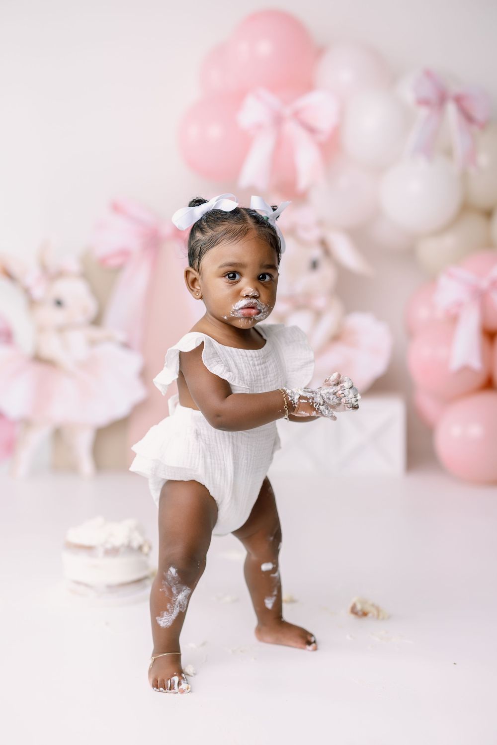 Baby girl in white outfit having fun during cake smash photoshoot with pink and white balloon backdrop.