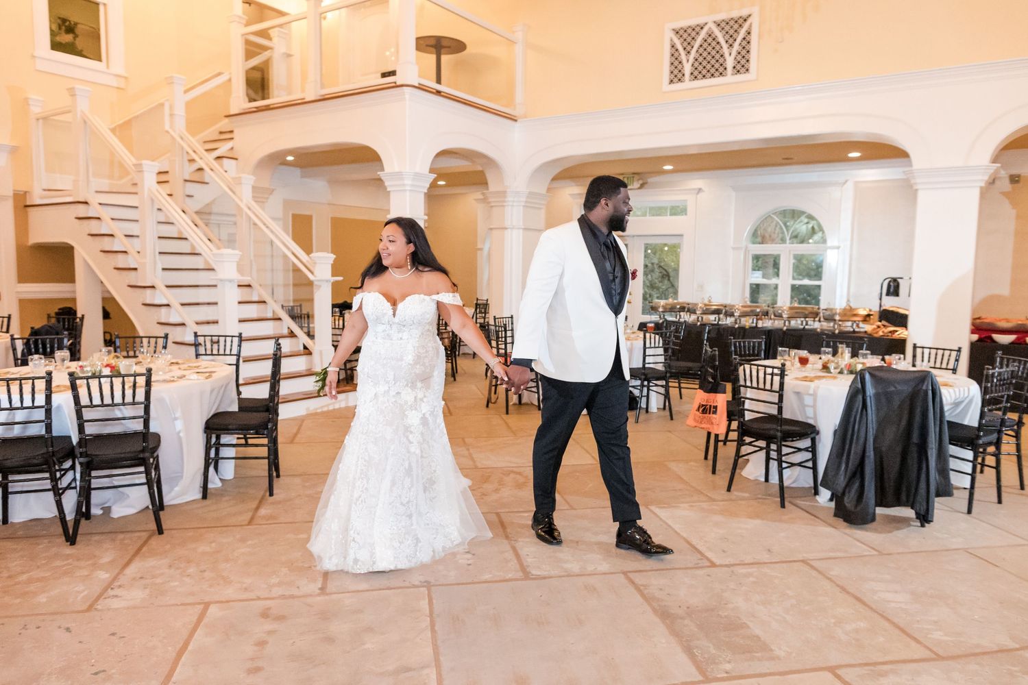 First look wedding photo of couple standing back to back in an elegant venue with white spiral staircase and arched windows.