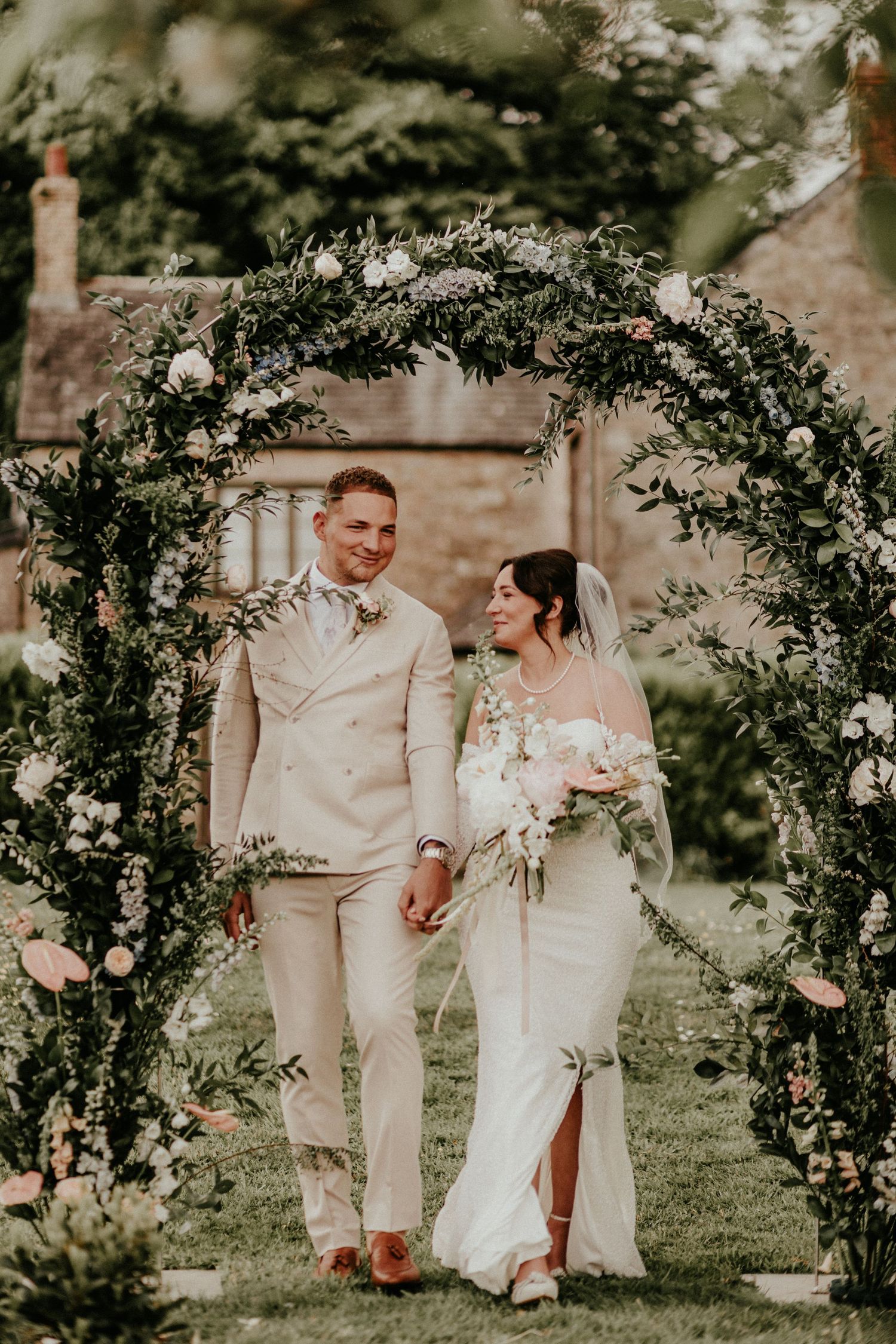 Couple walks through lush floral wedding arch with greenery and blush roses in an intimate garden ceremony.