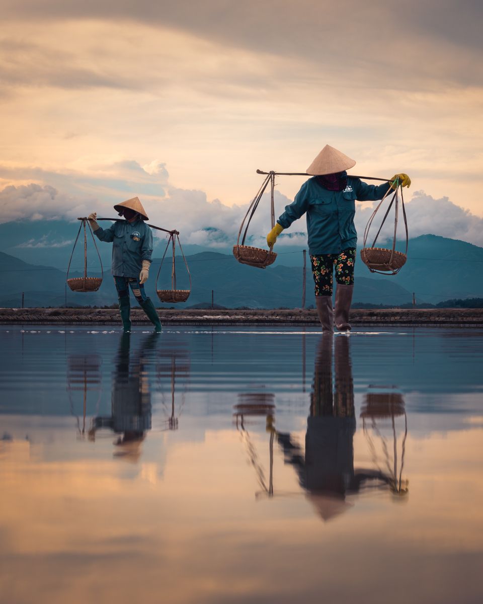 Workers carrying traditional yoke baskets harvest salt at sunset in Vietnam's salt fields with mountain reflections in water.