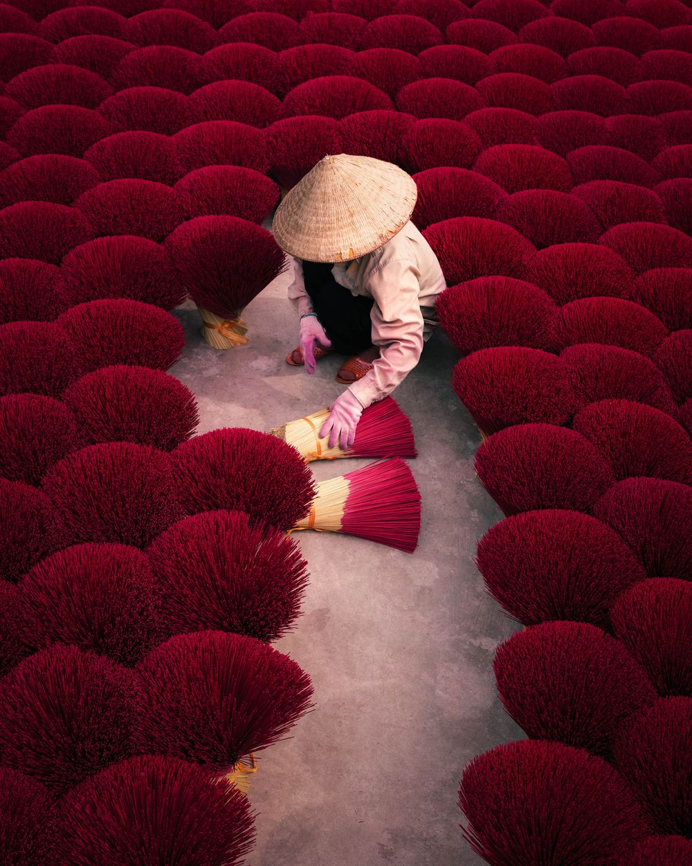Worker wearing traditional conical hat tends to vibrant red incense sticks drying in rows at a Vietnamese factory.