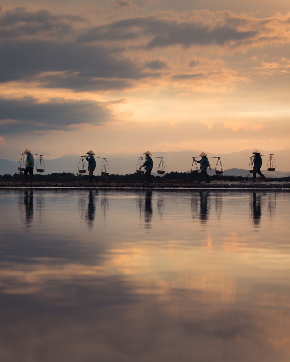 Silhouettes of salt farmers carrying shoulder poles are reflected in calm water during a dramatic sunset in Vietnam.