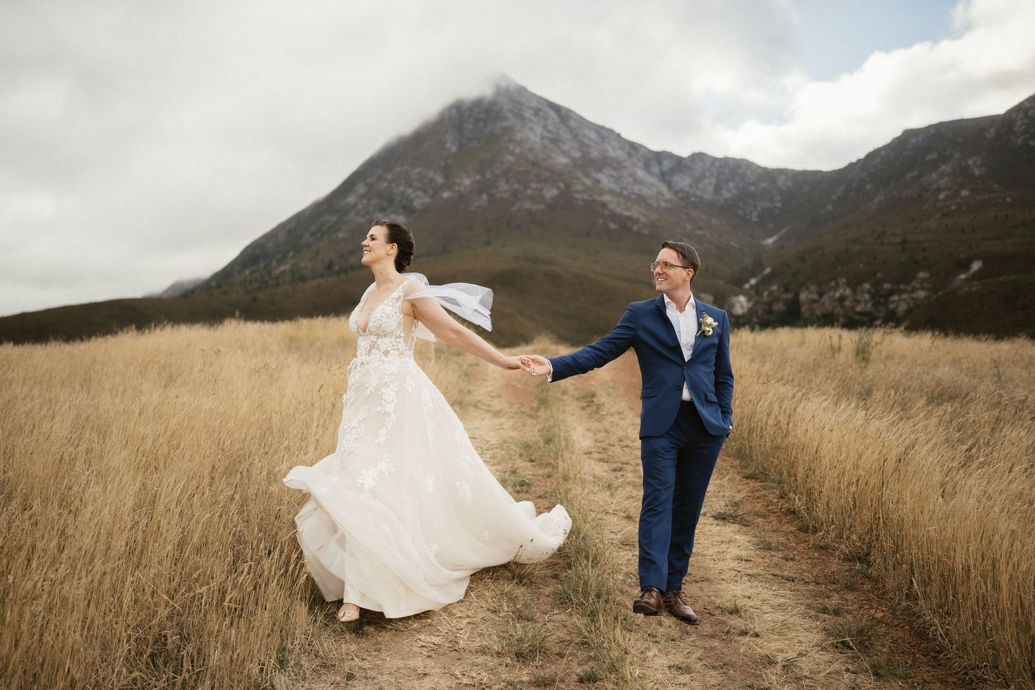 A couple holds hands while walking through tall golden grass with rugged mountain peaks in the background.
