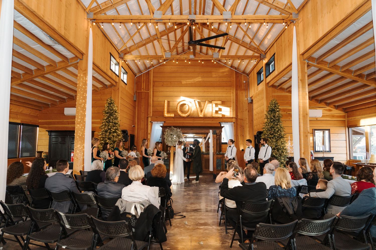 Wedding ceremony in a rustic wooden venue decorated with Christmas trees and illuminated LOVE sign.