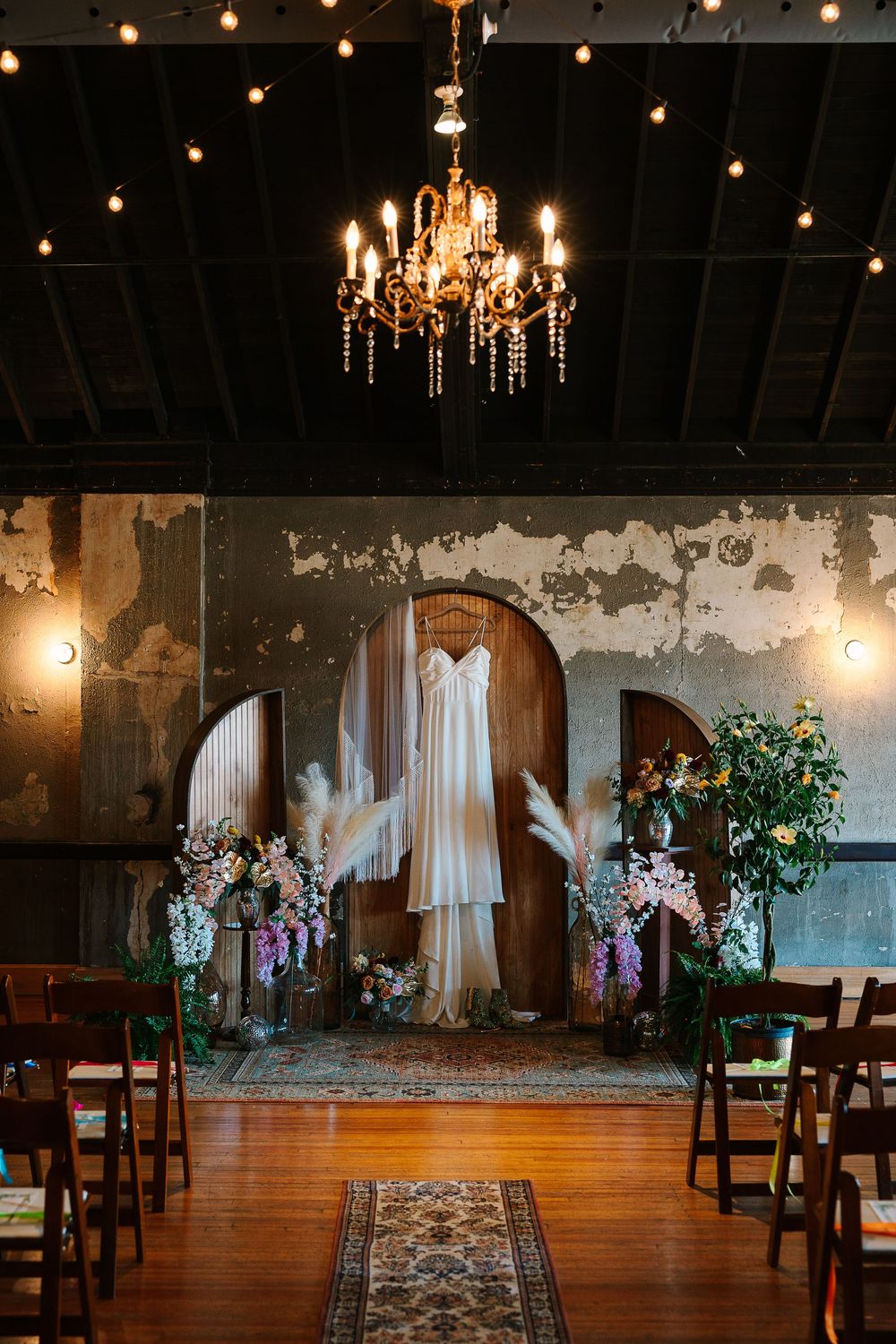A wedding dress with a fringe shawl hangs from a wooden arch, surrounded by eclectic flowers and ornate rugs with a chandelier overhead.