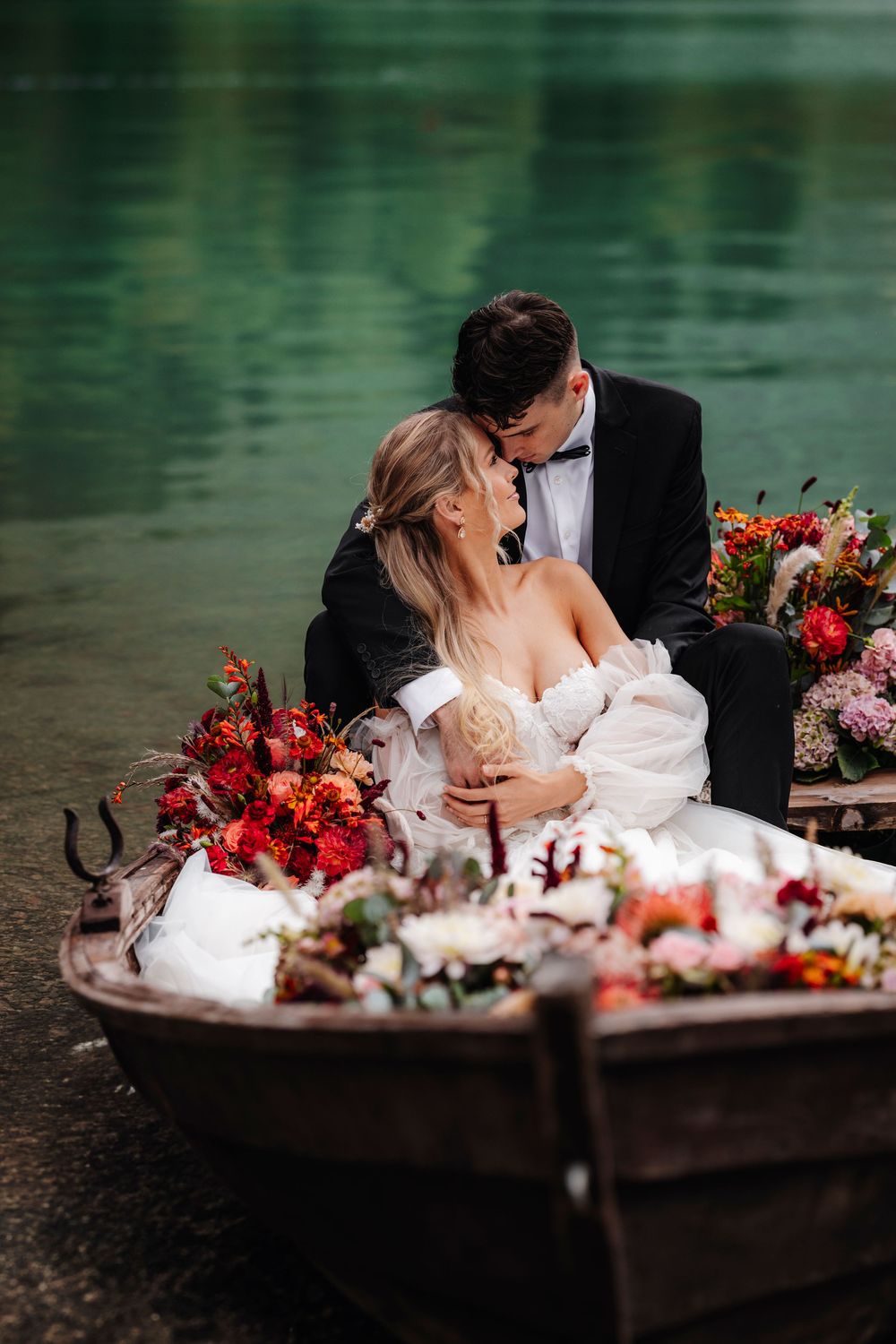 Romantic couple sharing an intimate moment in a boat filled with vibrant red and pink floral arrangements by a serene lake.